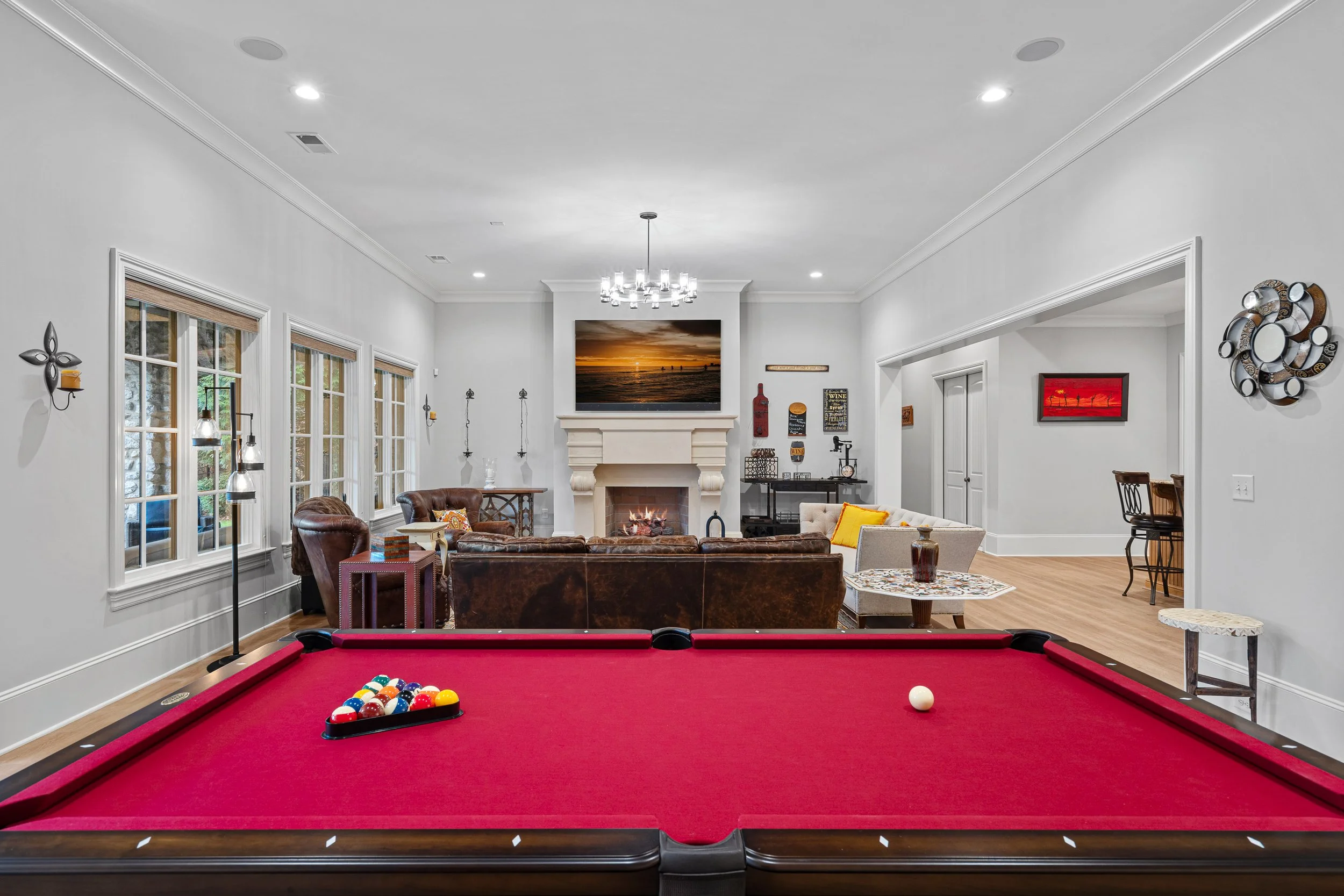 Interior of a living room with a red pool table in the foreground, leather and fabric sofas facing a fireplace and television, surrounded by large windows, modern light fixtures, and various wall and decorative art.