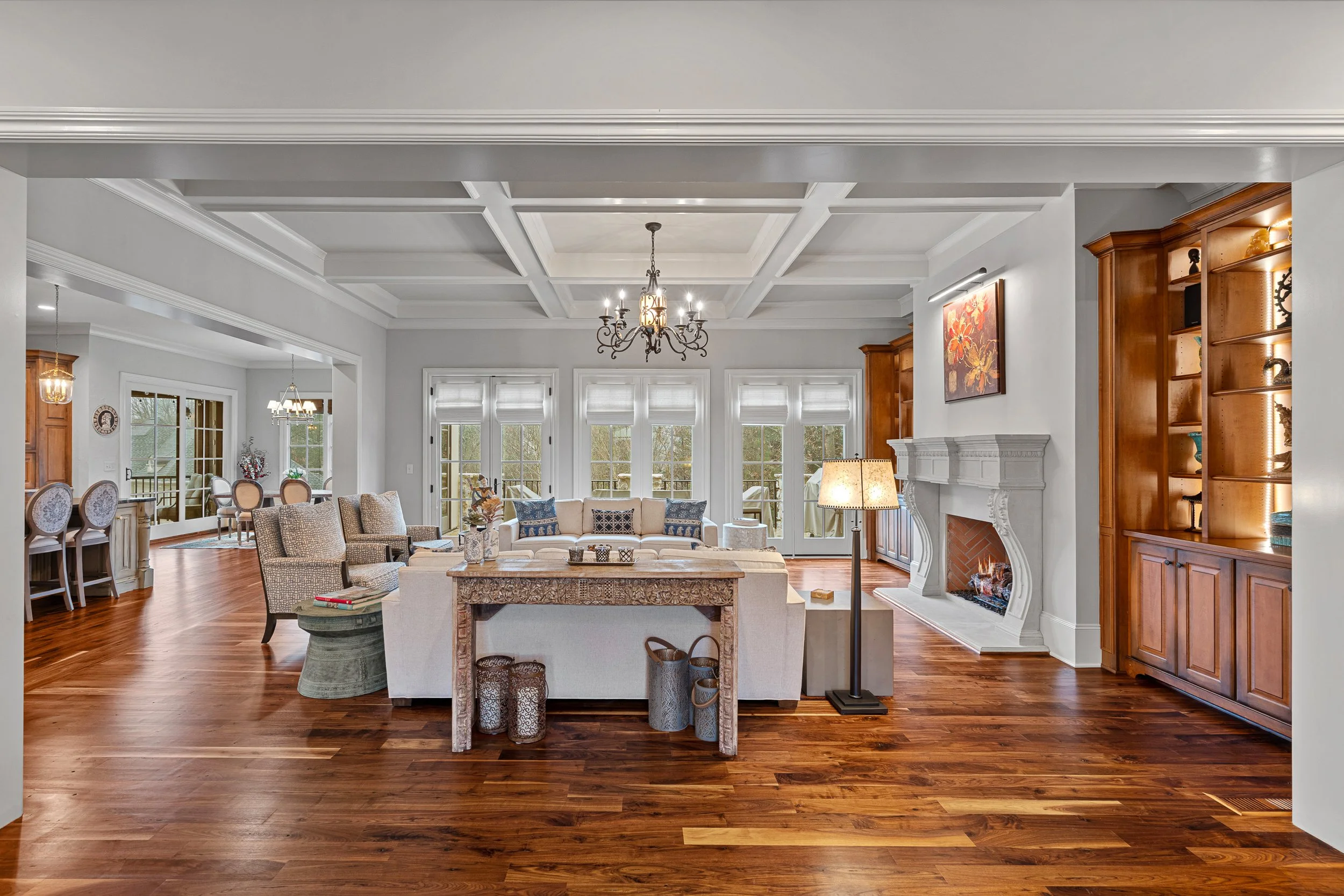 Spacious living room with white walls, hardwood floors, and large windows, featuring a white sofa, armchairs, a fireplace, and wooden built-in shelves.