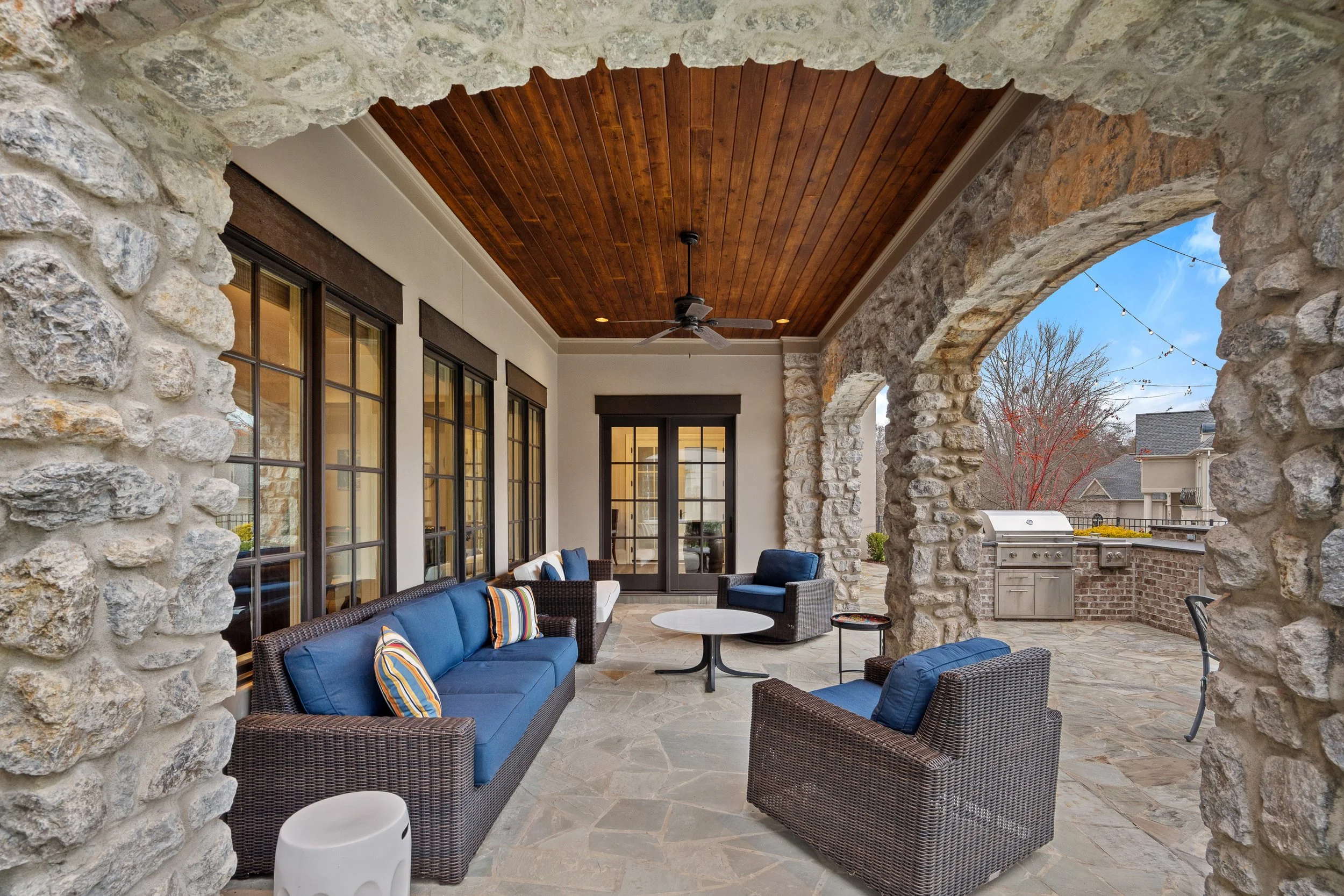Covered outdoor patio with stone arches, wicker seating with blue cushions, a round coffee table, and a ceiling fan, overlooking a backyard with a grill and string lights.