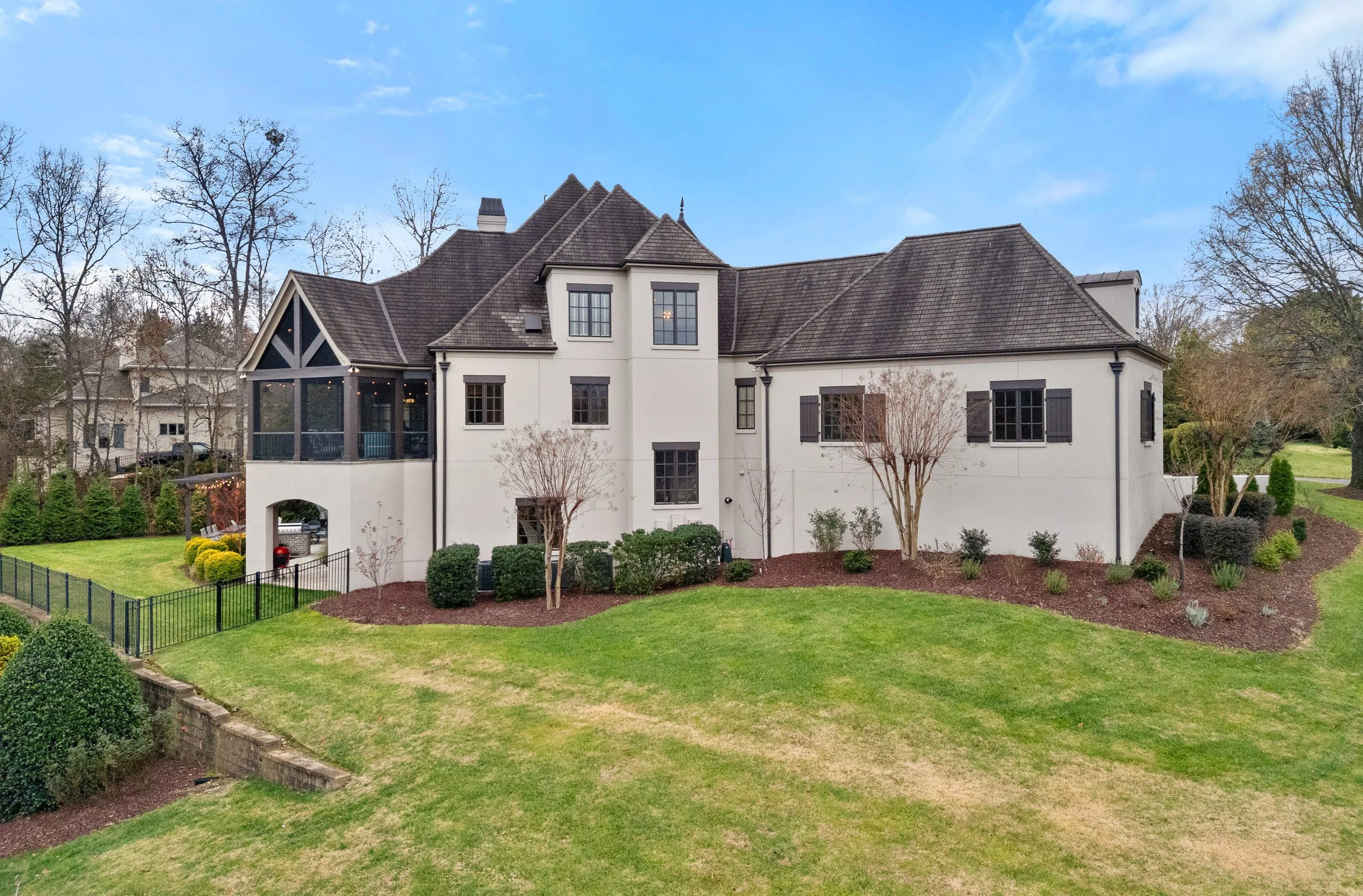 A large white house with a dark shingled roof, surrounded by a well-manicured lawn and trees, under a partly cloudy sky.
