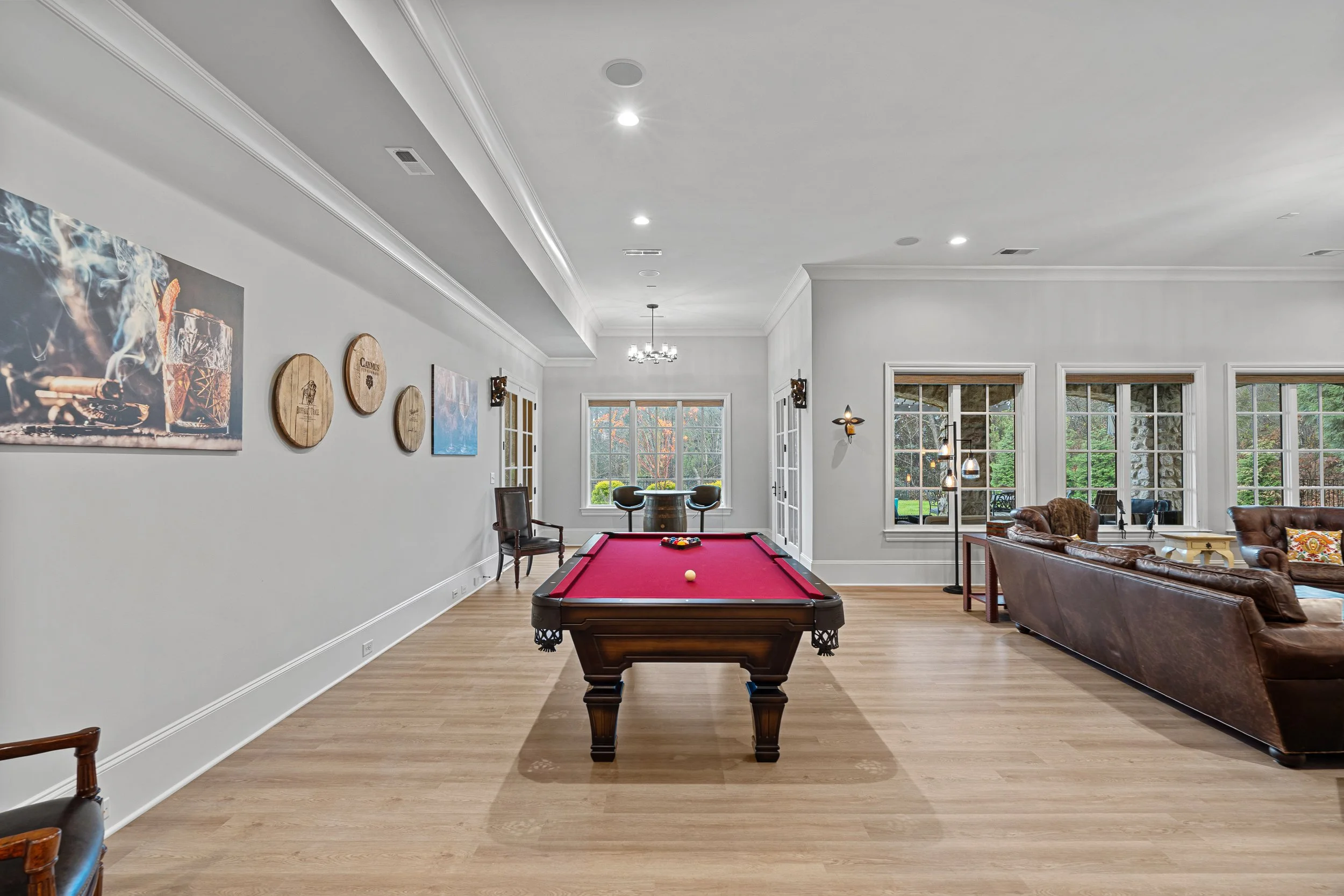 Living room with a red pool table in the center, brown leather sofa, art on the walls, and large windows with a view of greenery outside.