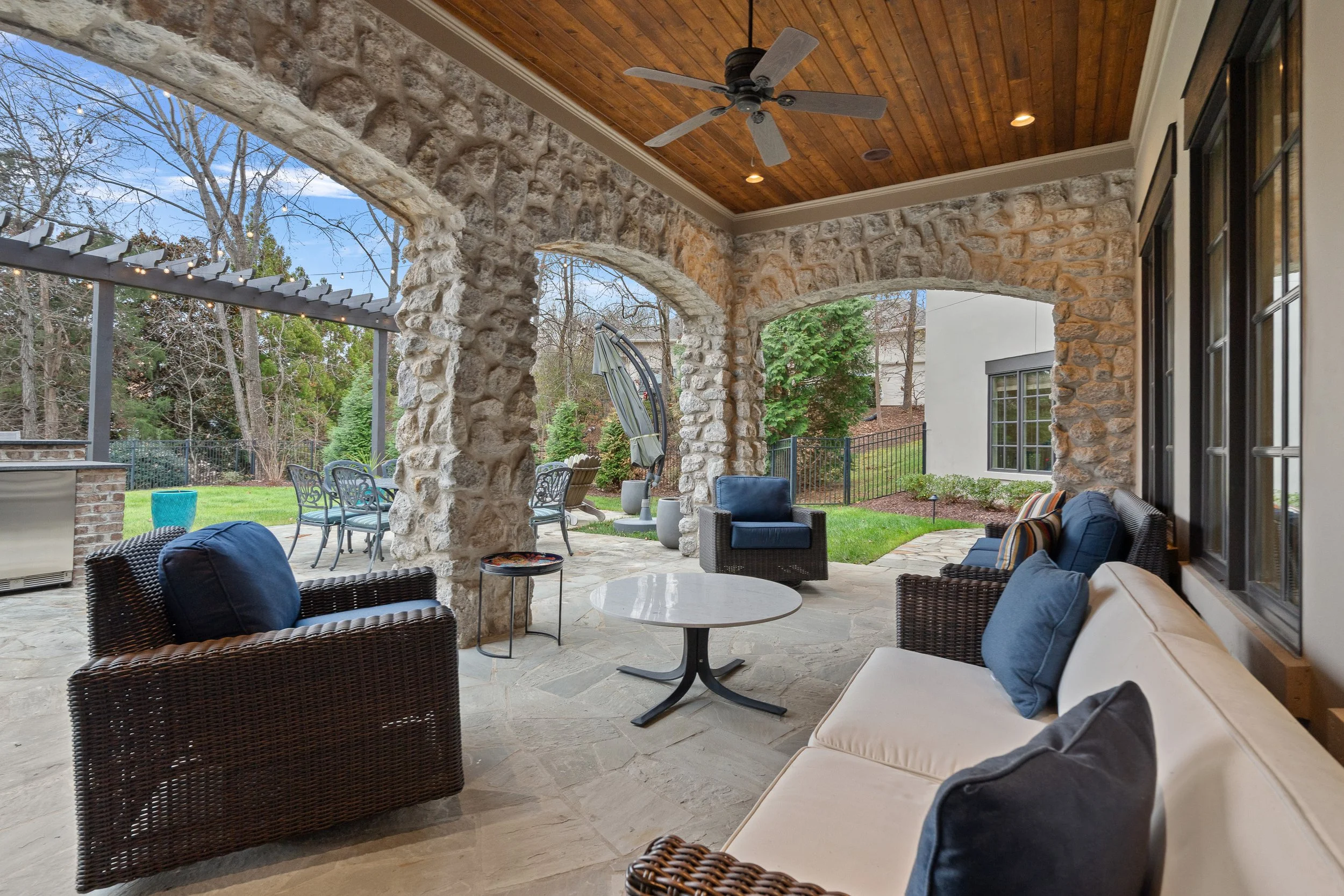 Covered outdoor patio with stone columns, wicker chairs with cushions, a marble-top table, and a ceiling fan, overlooking a backyard with trees, green grass, and outdoor furniture.
