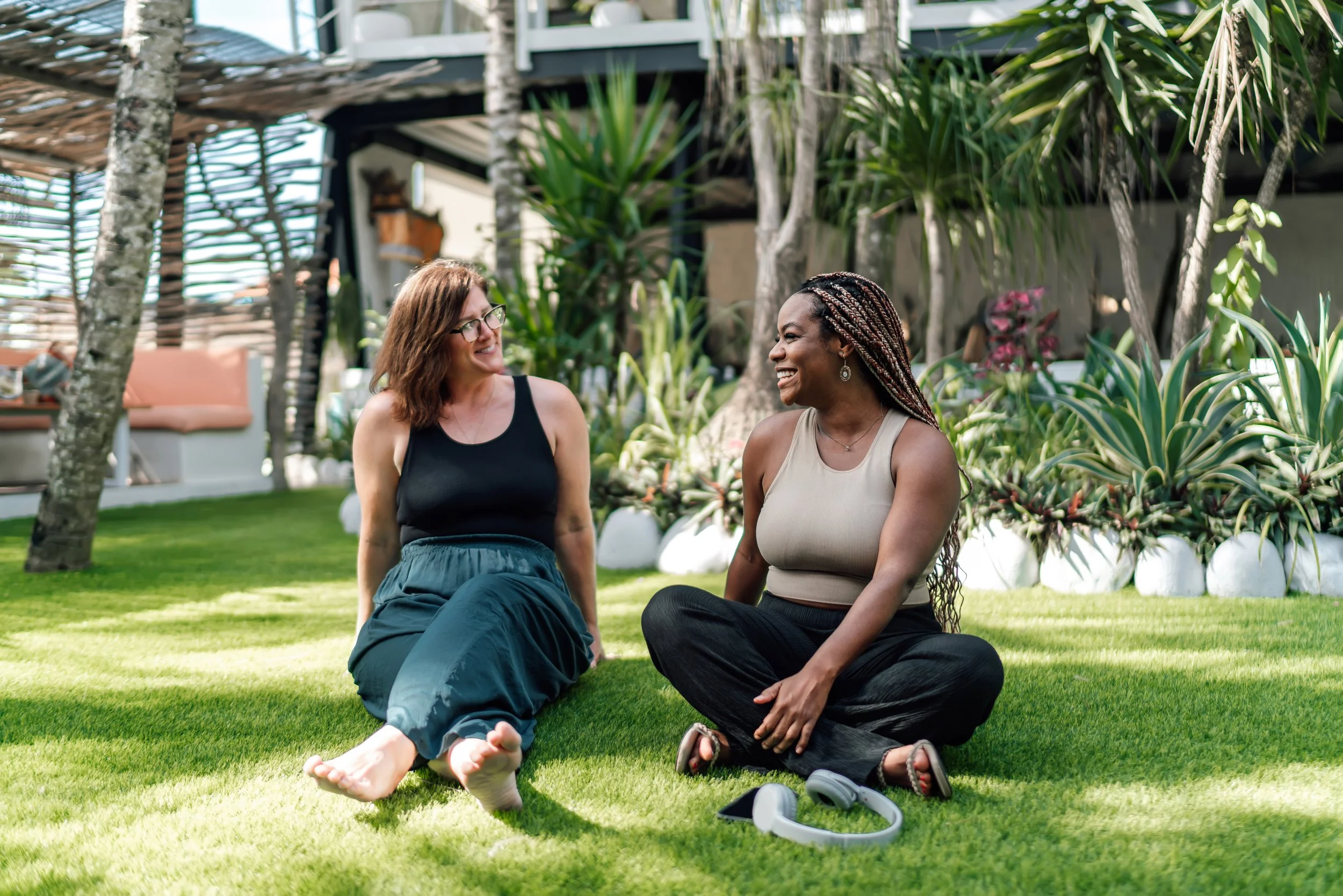 Two women sitting on the grass, smiling and talking in a lush garden with tropical plants.
