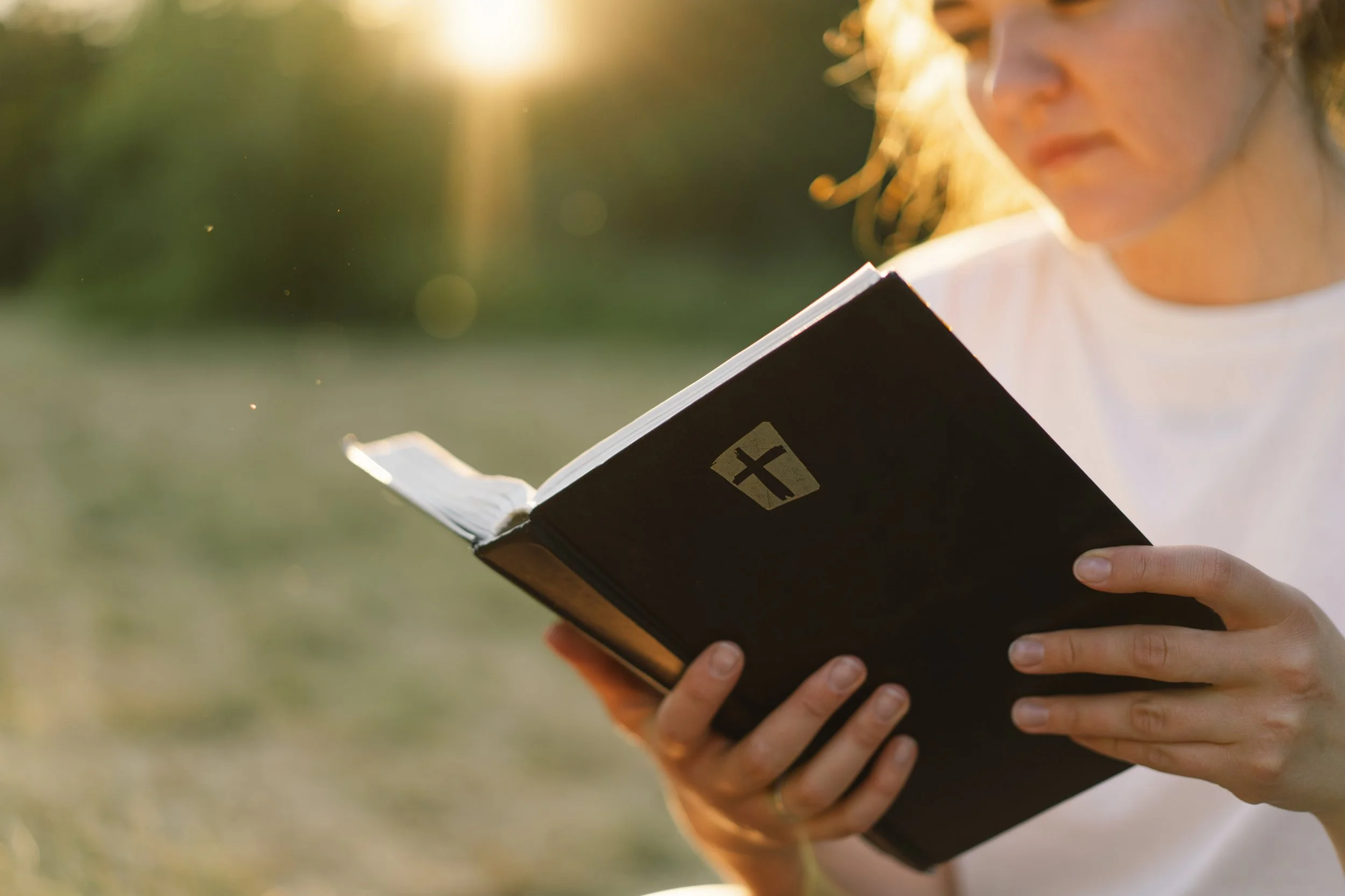 A woman reading a religious book outdoors in the sunlight, with a cross symbol on the cover.