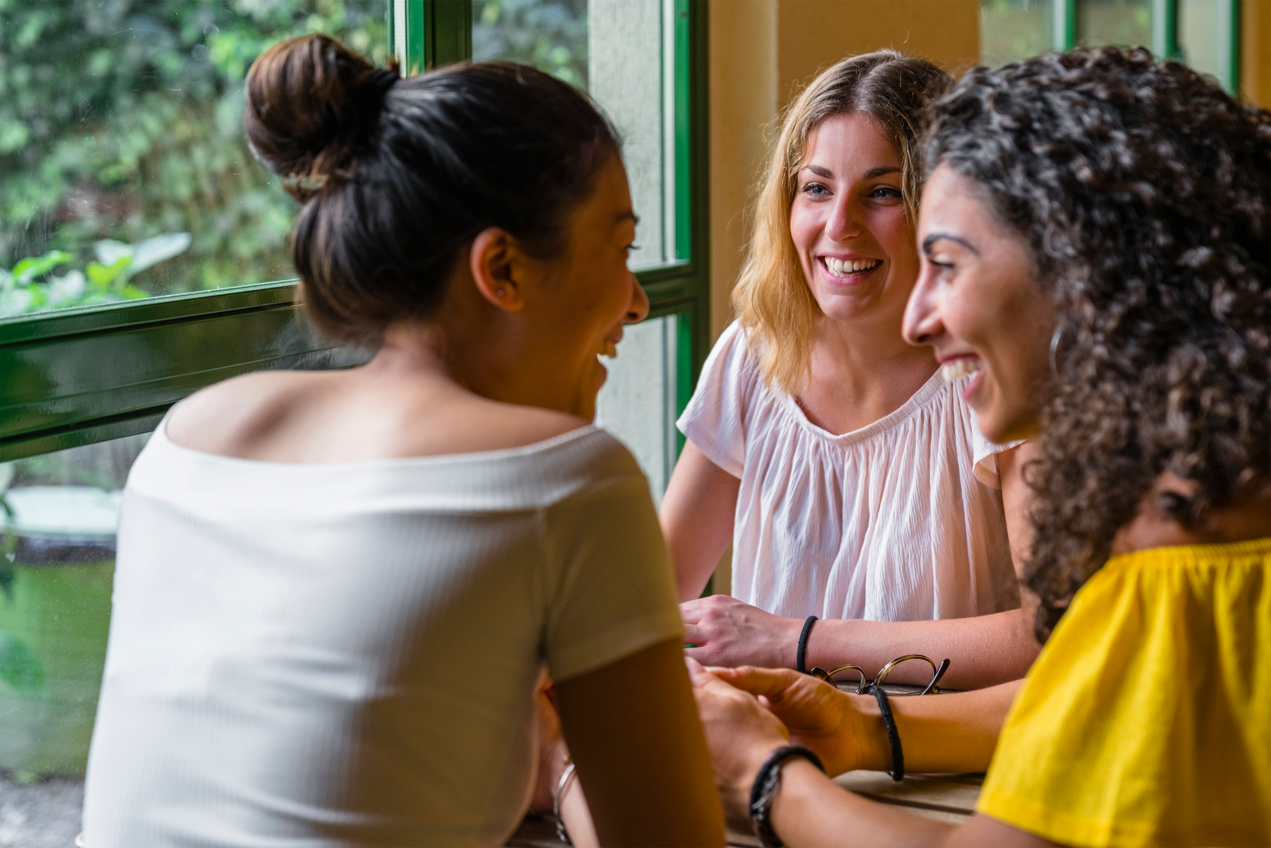 Four women sitting around a table, smiling and talking inside a cafe near a window.
