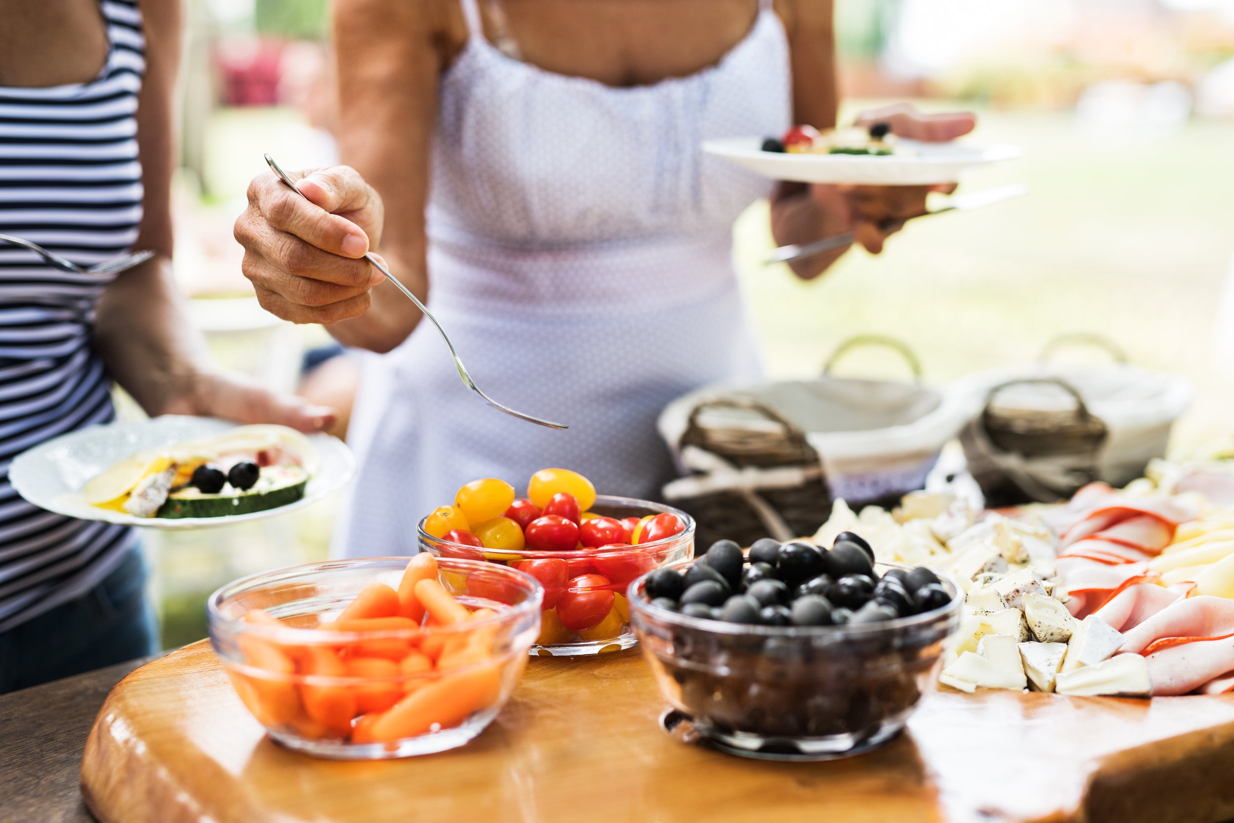 People serving themselves food at a buffet table with bowls of cherry tomatoes, baby carrots, and cheeses.