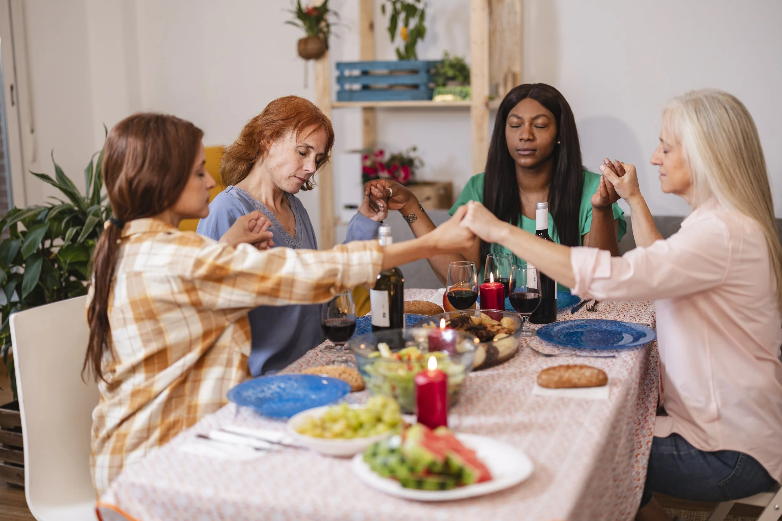 Four women praying at a dinner table decorated with candles, wine, and food, holding hands with eyes closed.