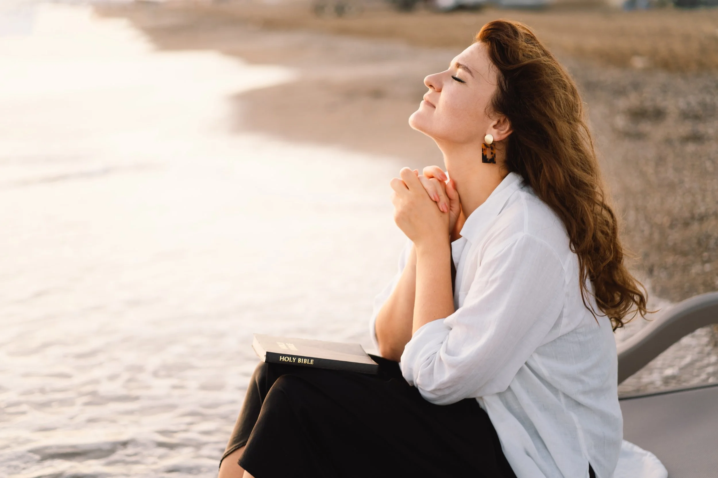 A woman sitting on a beach with her eyes closed, praying or meditating, holding a Bible on her lap, wearing a white shirt and large earrings, during sunset.