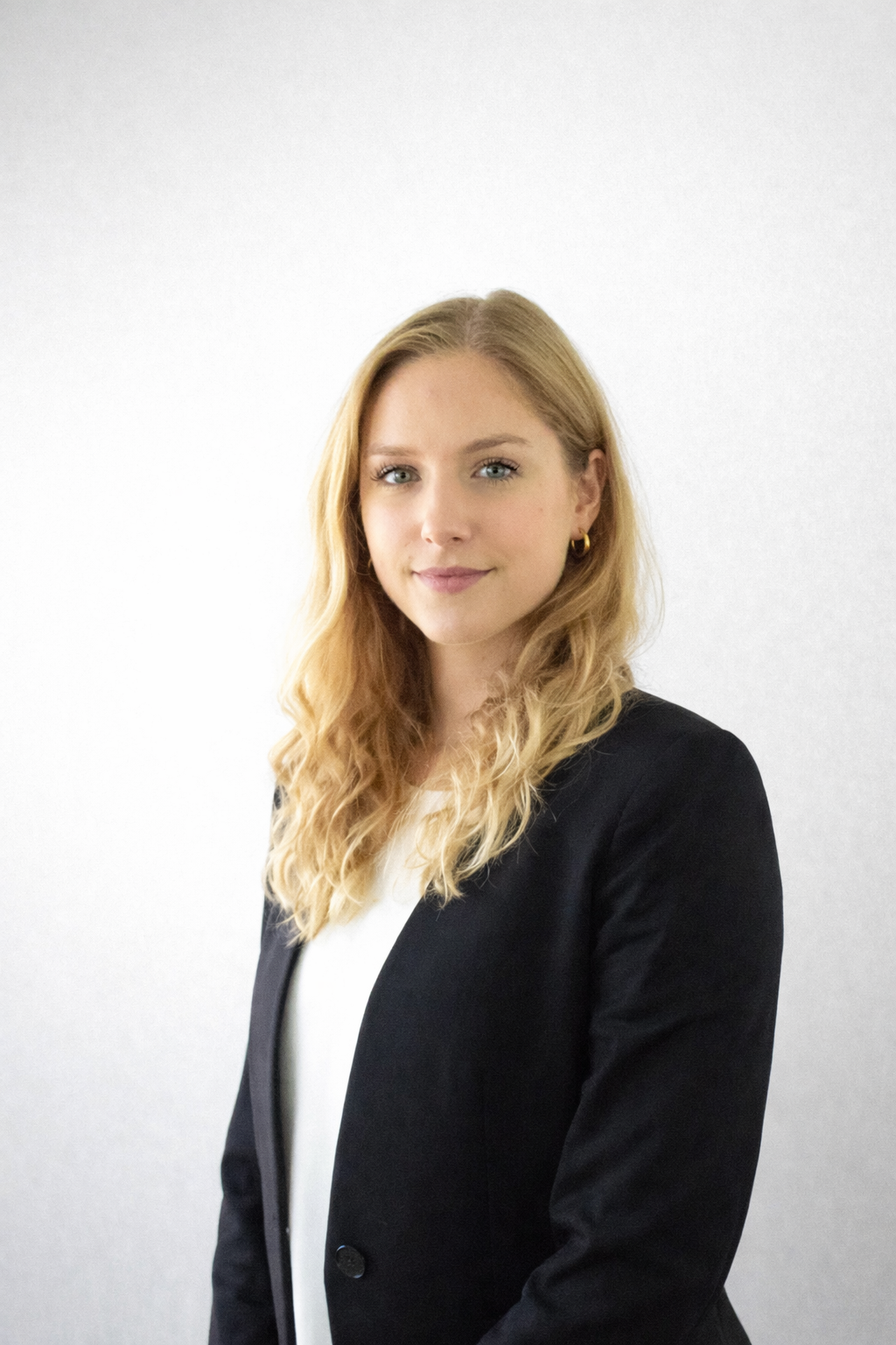 Johanna Ruwolt - Porträt einer jungen Frau mit blonden, gewellten Haaren, blauen Augen und einem freundlichen Lächeln, trägt einen schwarzen Blazer und eine weiße Bluse vor einer weißen Wand.