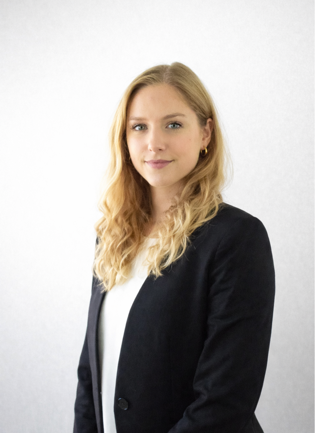 Johanna Ruwolt - Porträt einer jungen Frau mit blonden, gewellten Haaren, blauen Augen und einem freundlichen Lächeln, trägt eine schwarze Blazer und eine weiße Bluse vor einer weißen Wand.