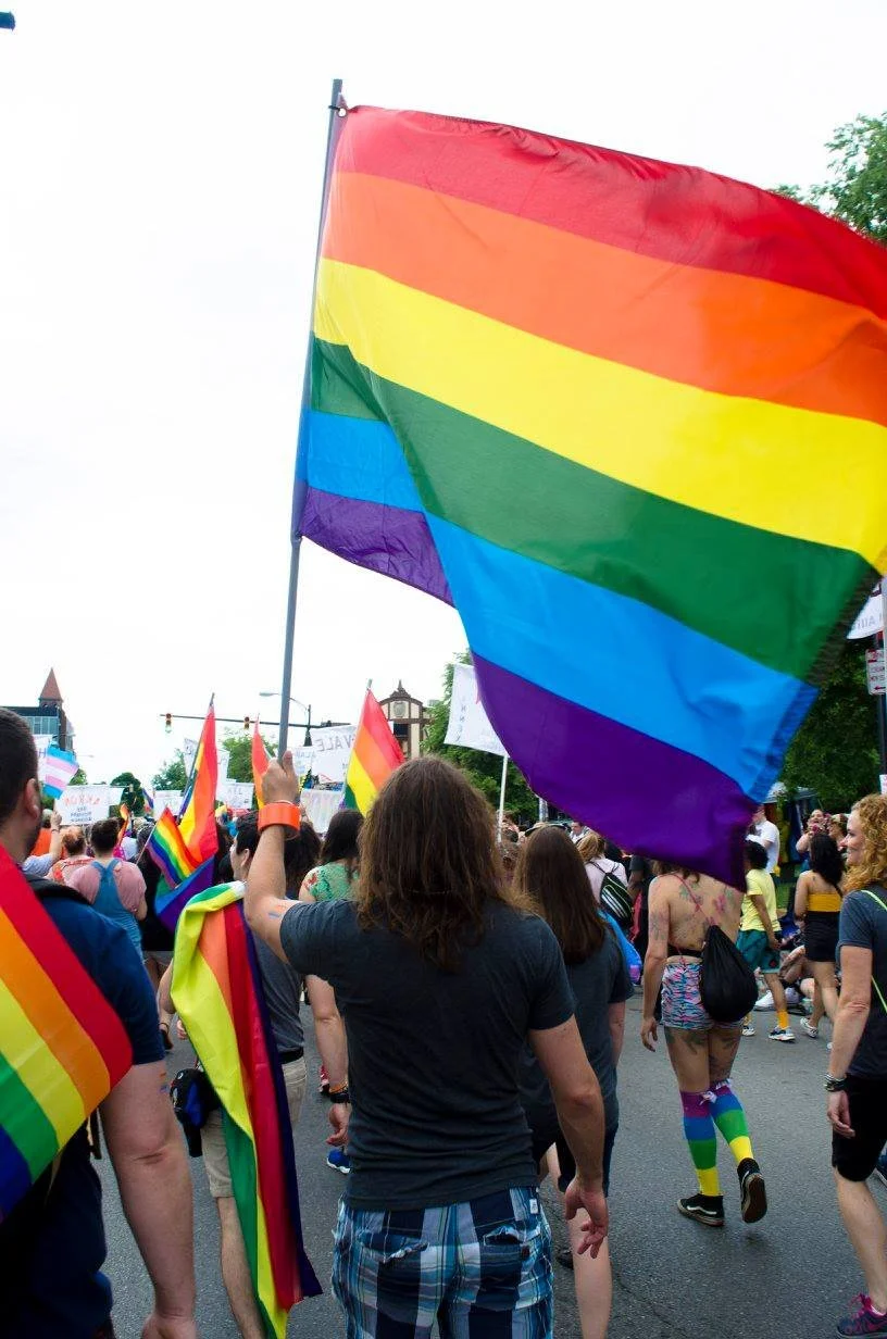People participating in a pride parade, holding rainbow flags and wearing rainbow-themed clothing.