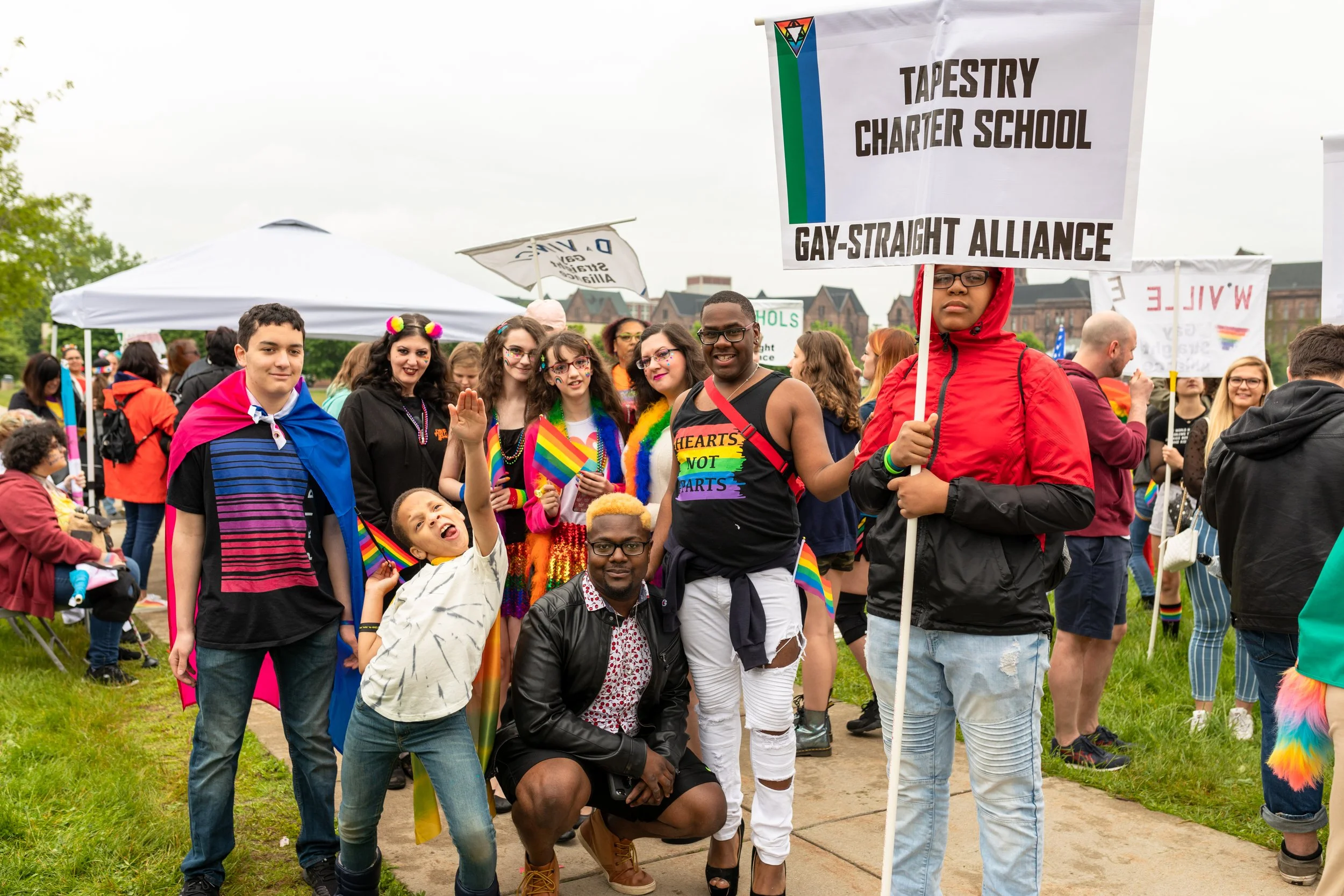 Group of people at a Pride parade holding LGBTQ+ pride flags and signs, with a tent and trees in the background.