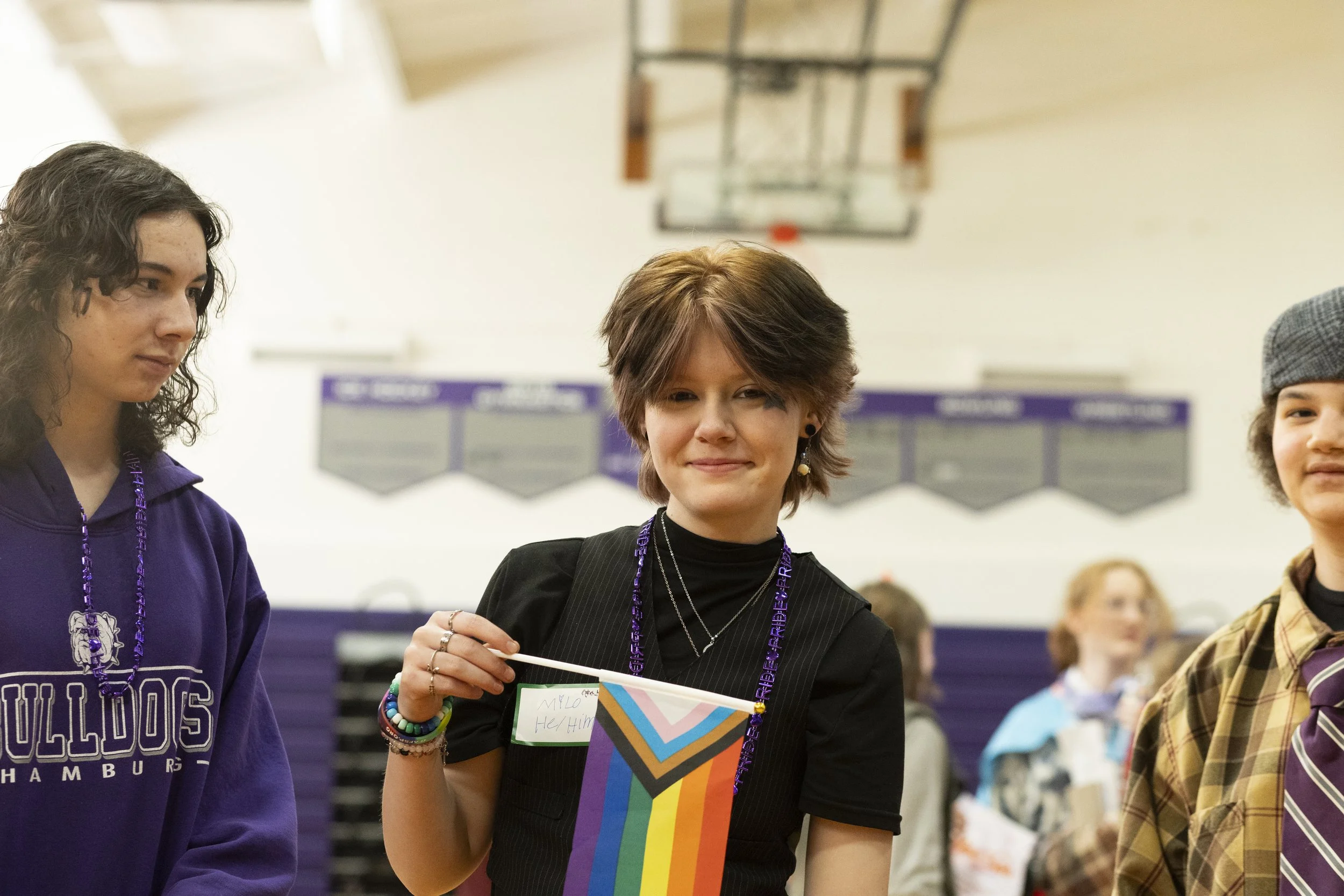 Young person at school gym holding rainbow pride flag, smiling, wearing black shirt and layered necklaces, with others in the background.
