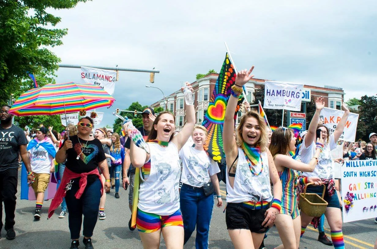 People celebrating at a Pride parade holding rainbow flags and signage, wearing colorful clothing and accessories, smiling and raising their hands.