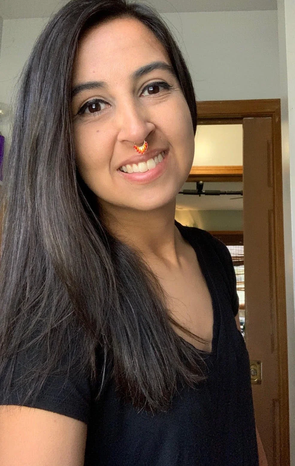 A woman with long dark hair smiling, wearing a black shirt, standing inside a house near a doorway.