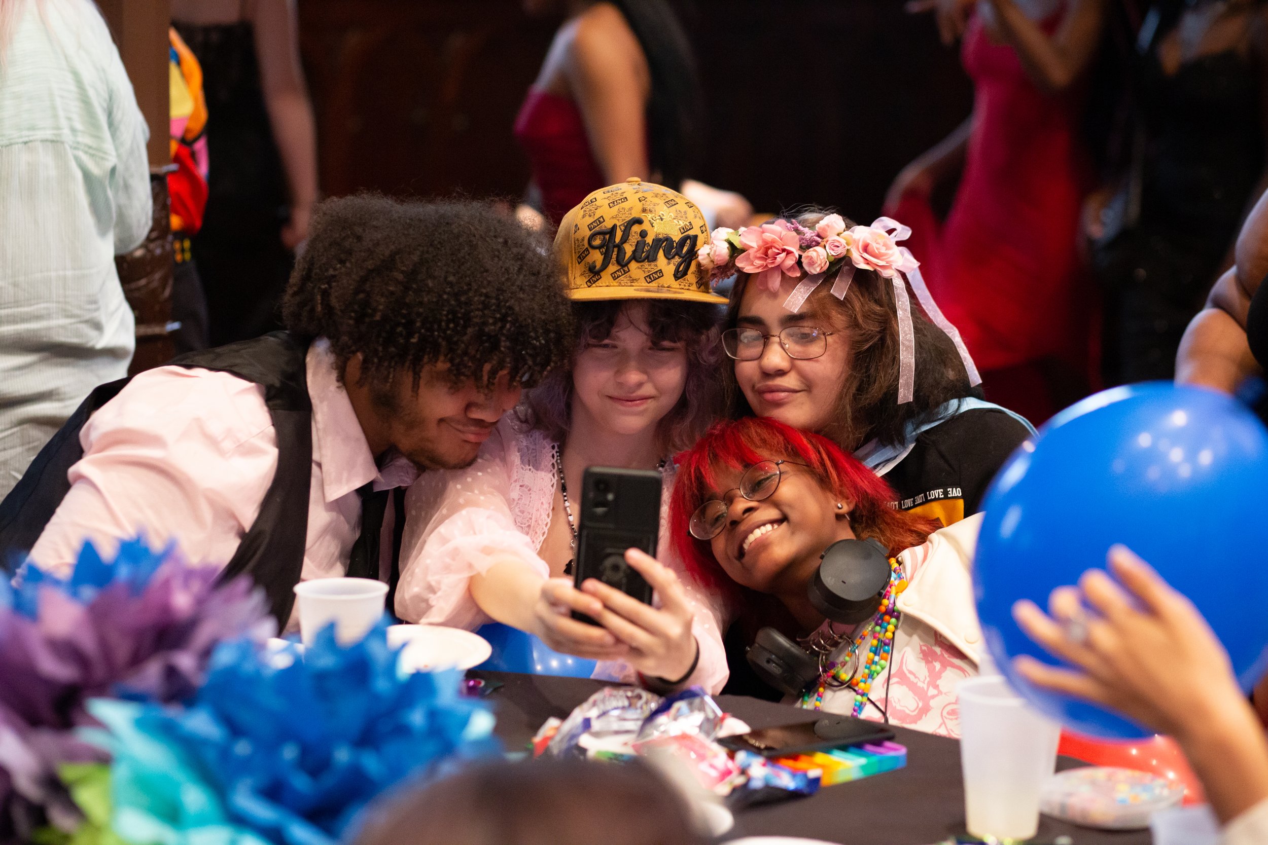 Four young people, three girls and one boy, are taking a selfie together at a celebration. They are sitting at a table with balloons, cups, and decorations in front of them. The girls are wearing colorful accessories, and one has a floral headband. The boy has curly hair and is smiling. All of them are smiling and appear happy.