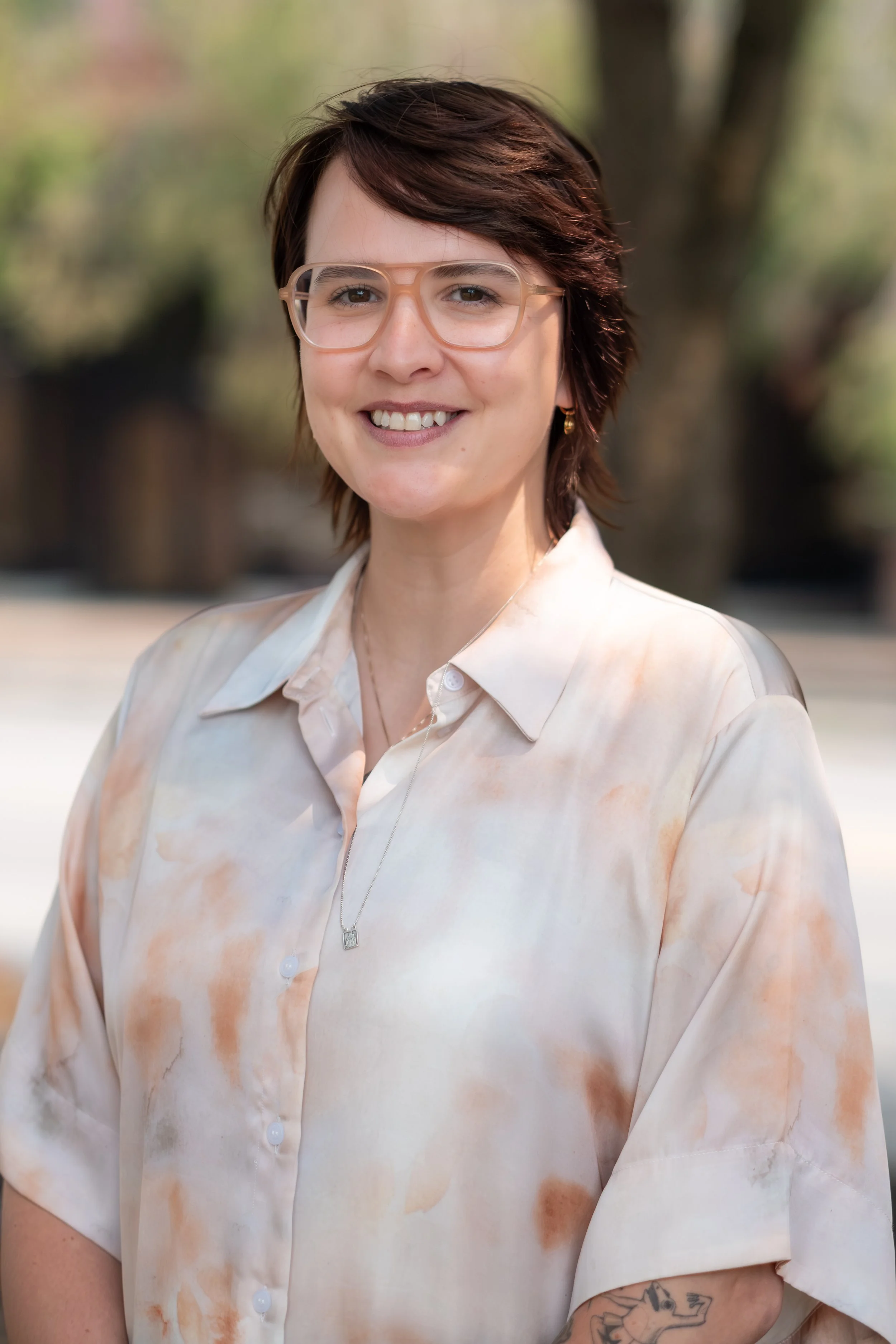 A person with short brown hair and glasses, smiling outdoors, wearing a light-colored, patterned button-up shirt.