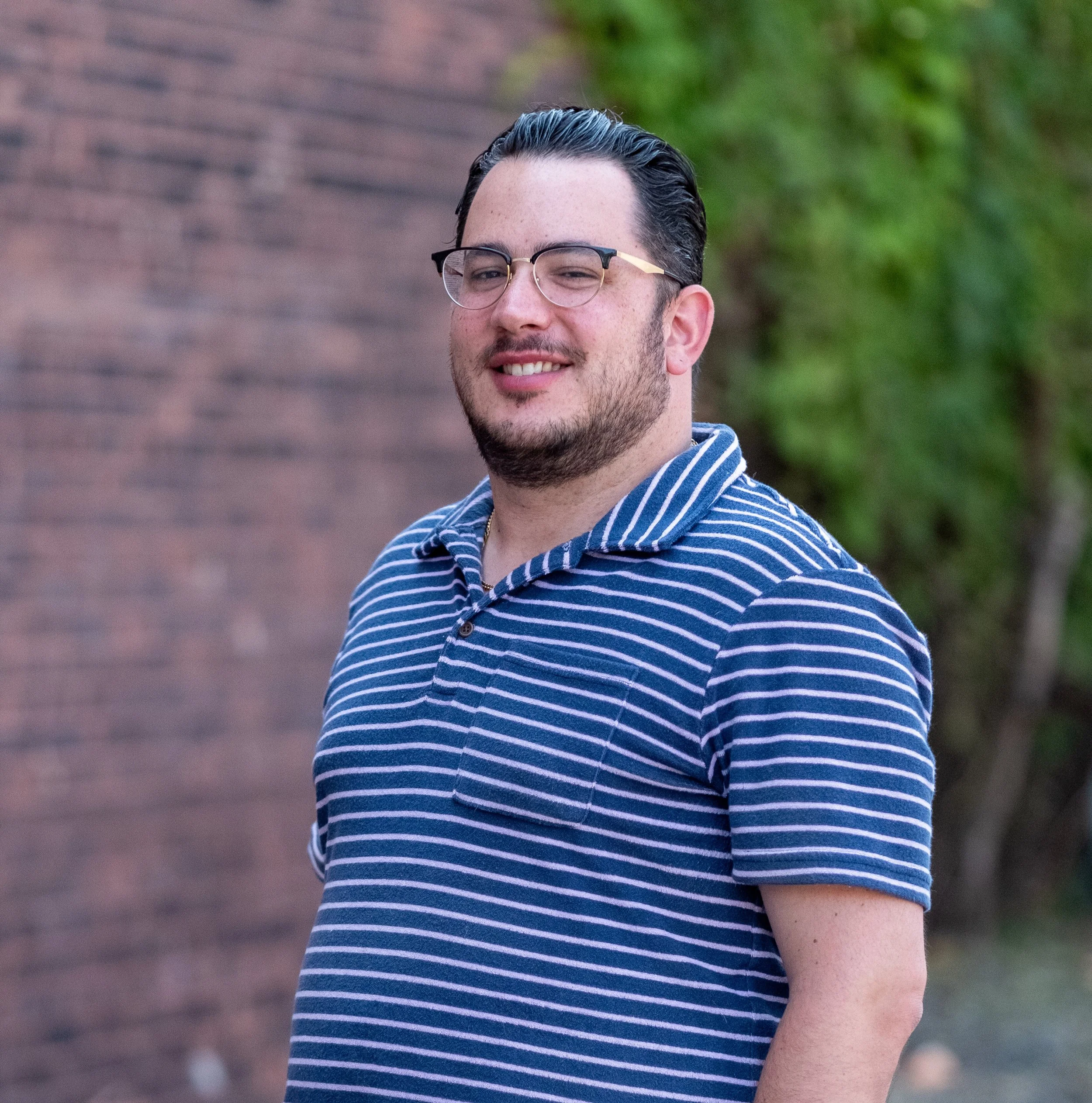 A young man wearing glasses and a blue and white striped polo shirt, smiling outdoors near a brick wall and greenery.