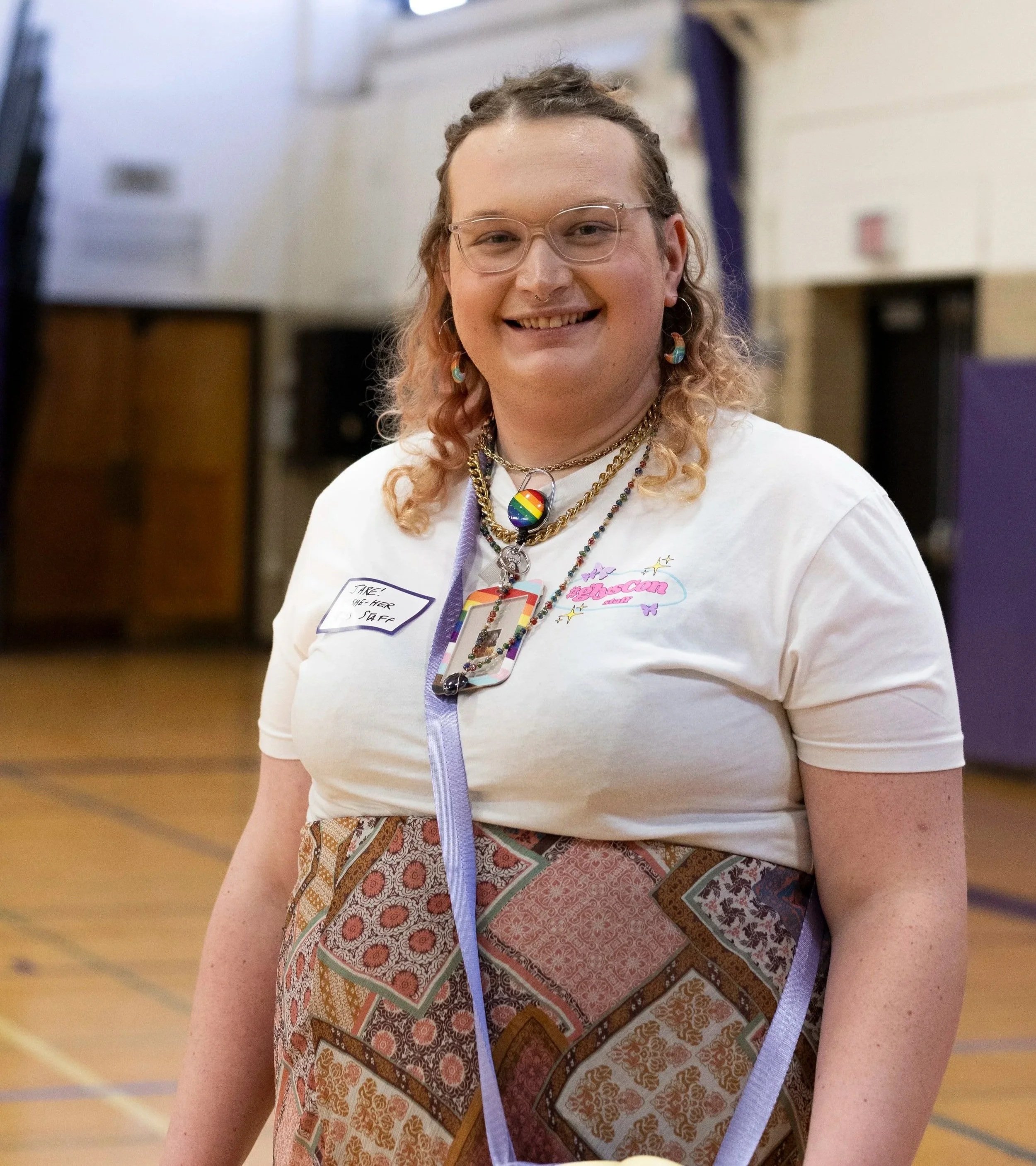 A young woman with curly hair, glasses, and a blue bandana standing in front of a brick wall. She is wearing a striped shirt, floral skirt, and layered necklaces, with colorful bracelets on her wrist.