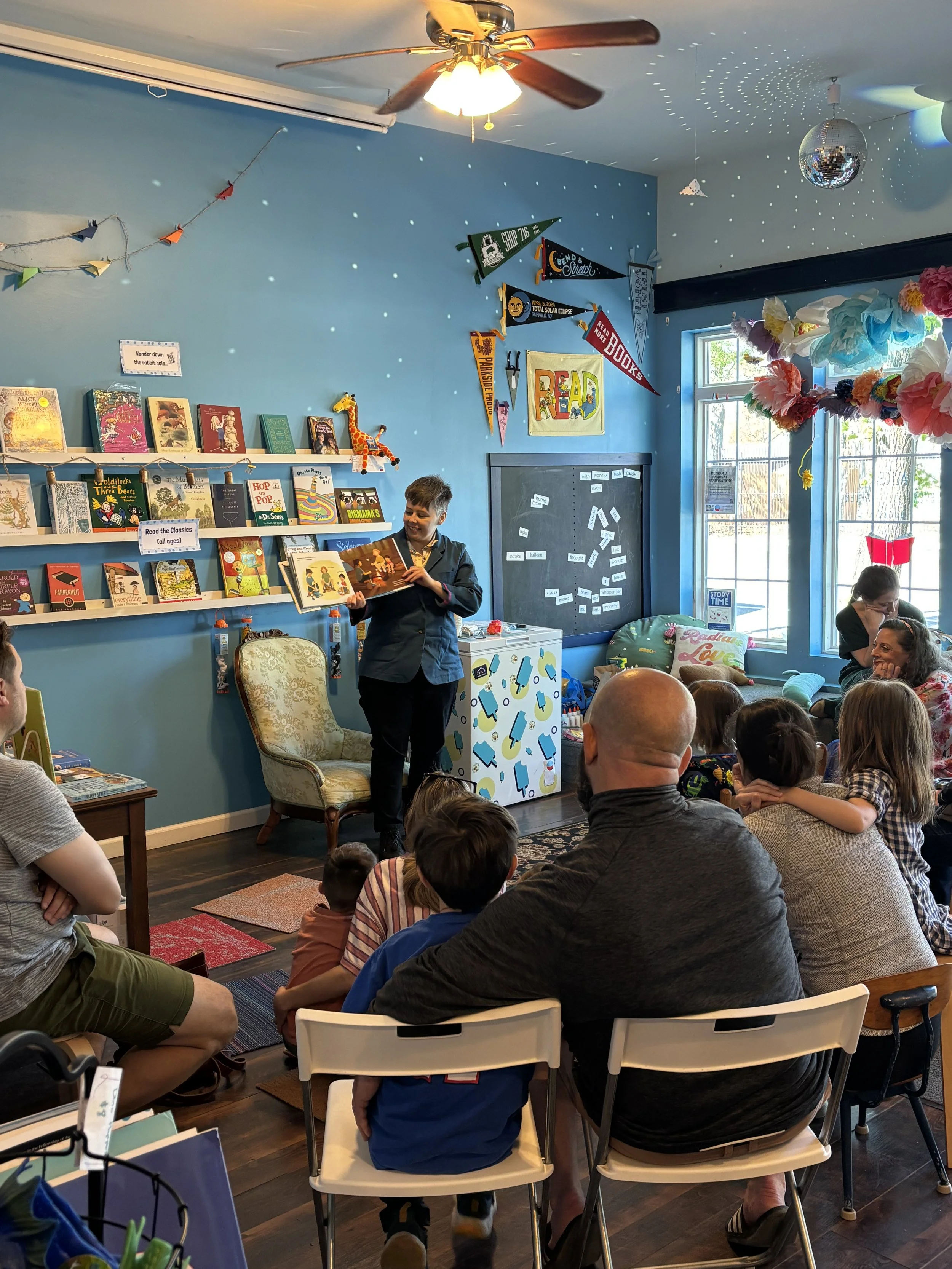 A person standing in front of a small audience, reading a book during a storytime event in a colorful, decorated room. The room has bookshelves, banners, and decorations including a disco ball and tissue paper flowers.