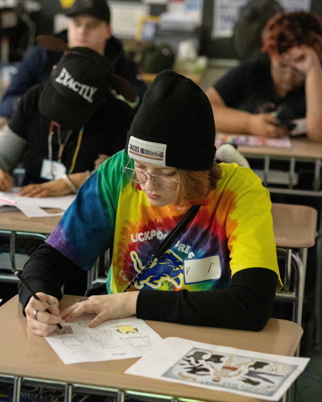 A person in a colorful tie-dye shirt and black beanie writing on paper at a desk in a classroom or workshop, with other people in the background.