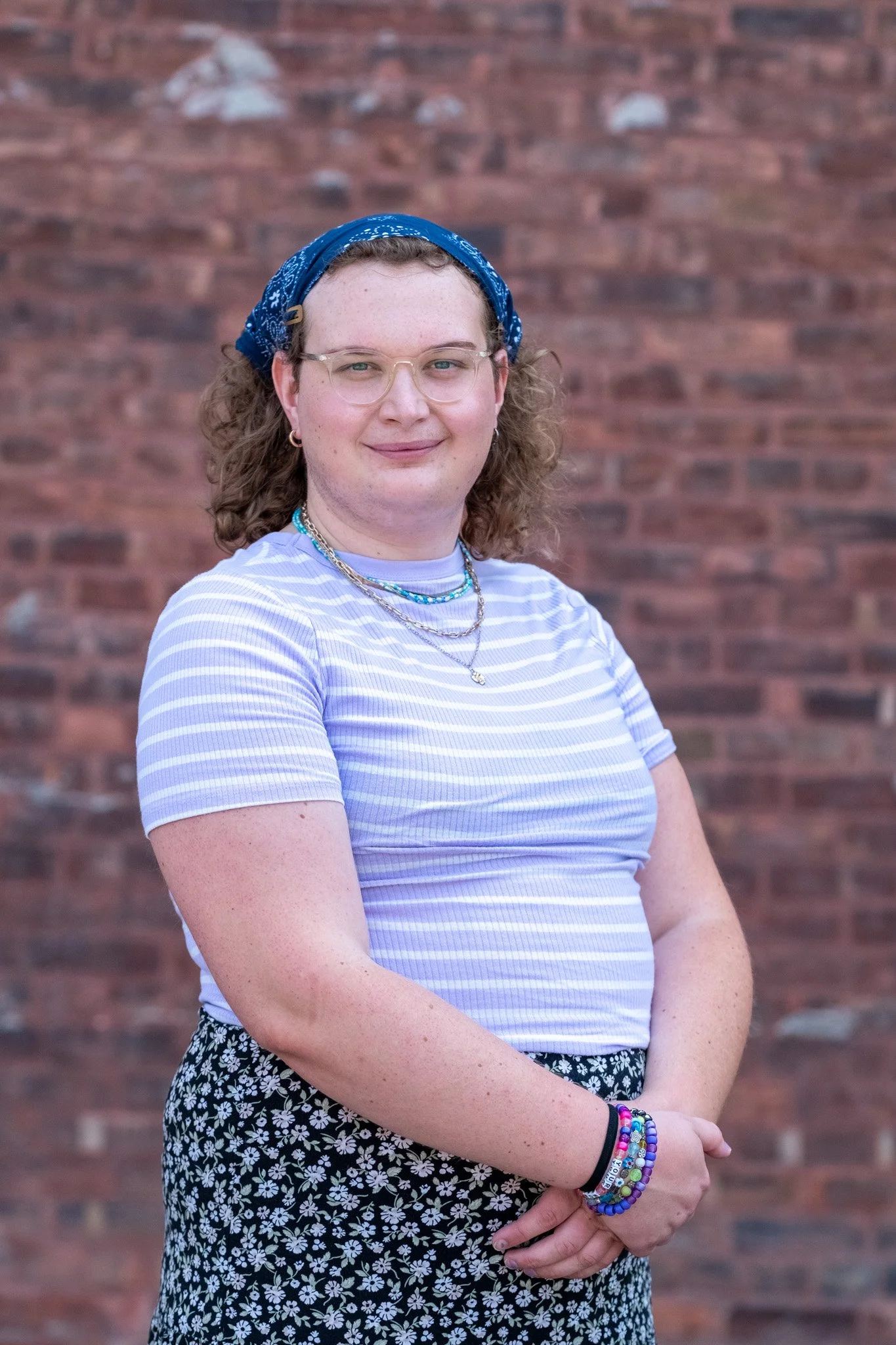 A young woman with curly hair, glasses, and a blue bandana standing in front of a brick wall. She is wearing a striped shirt, floral skirt, and layered necklaces, with colorful bracelets on her wrist.
