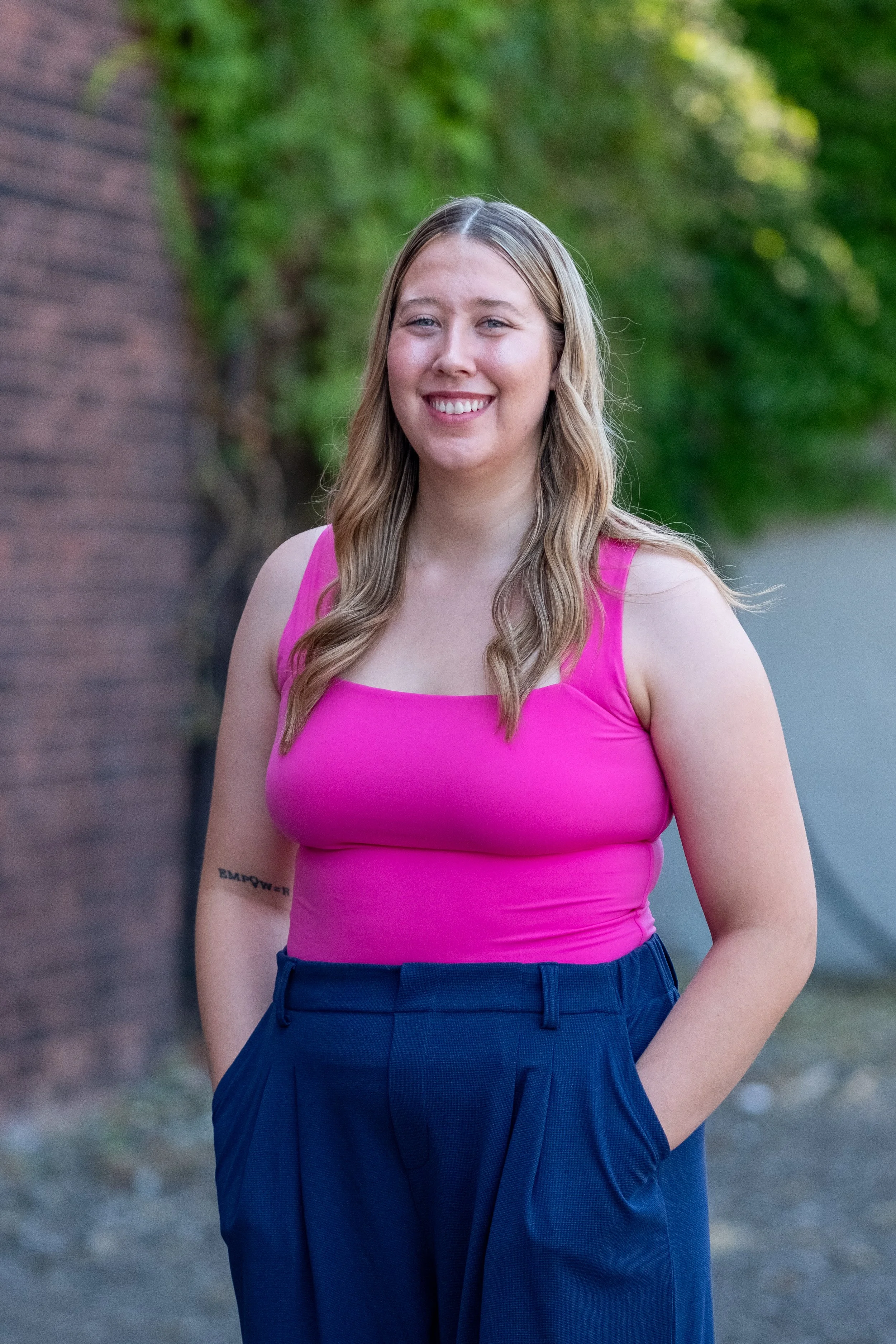A woman with long, wavy blonde hair wearing a bright pink tank top and high-waisted navy blue pants, smiling outdoors with greenery and a brick wall in the background.