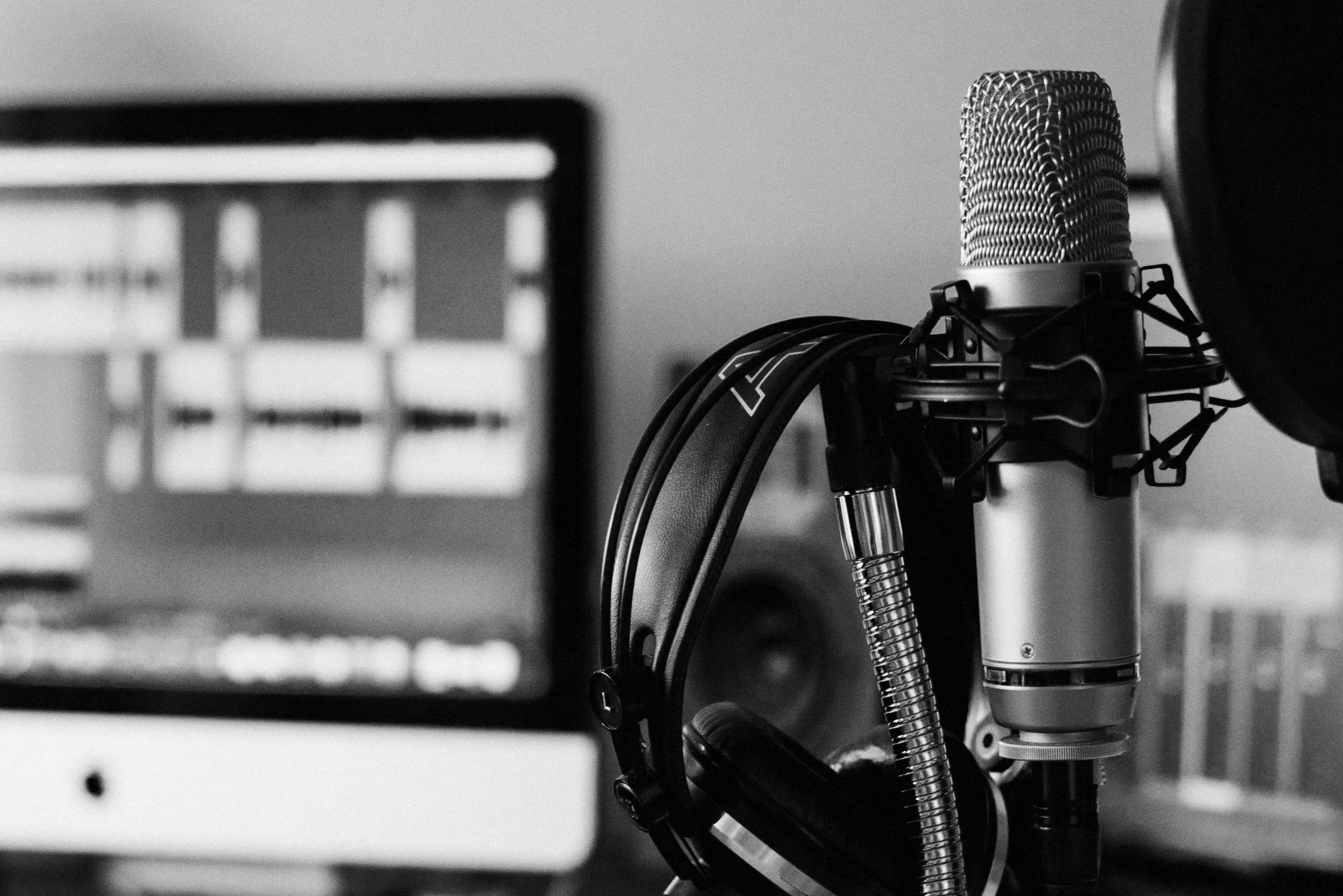 Close-up of a professional microphone with a pop filter and headphones in front of a computer monitor, suggesting a recording or broadcasting studio in black and white.