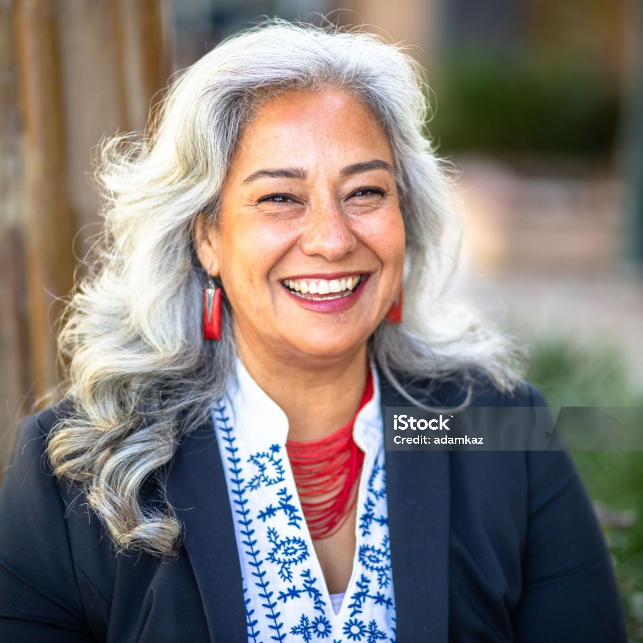 A smiling woman with long gray hair, wearing red earrings, red necklace, and a black blazer, sitting outdoors.