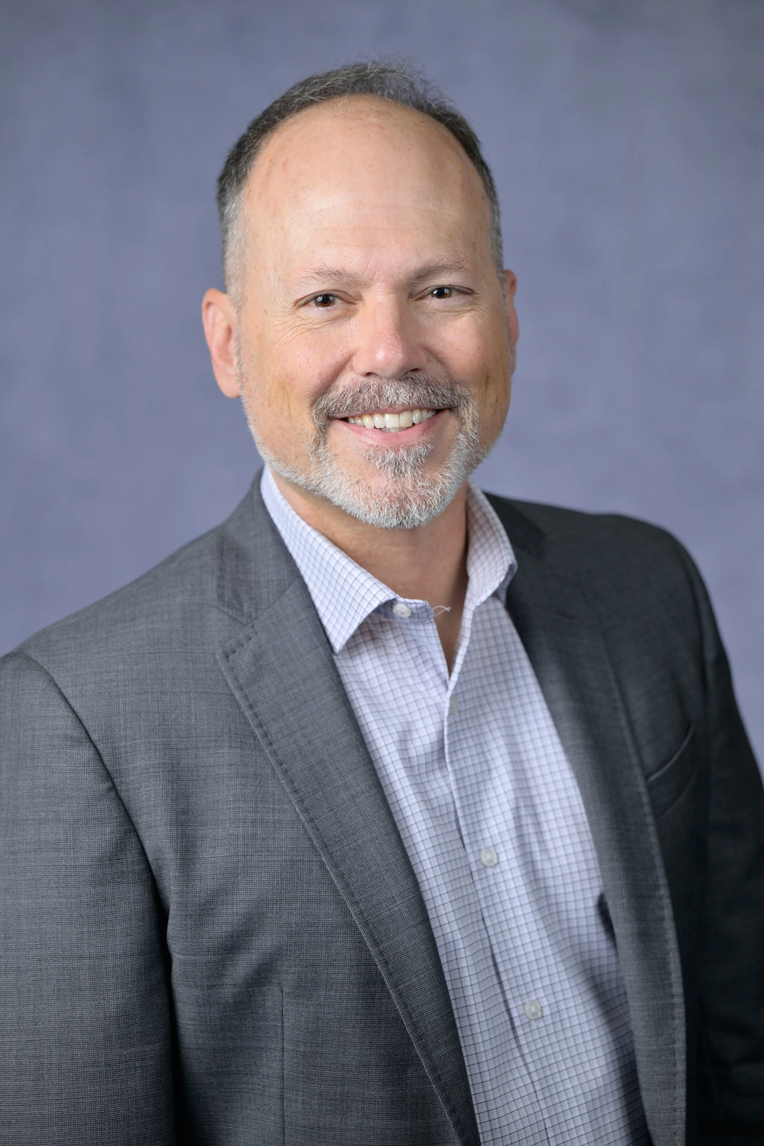 A middle-aged man with a beard and short hair, dressed in a gray business suit and checkered shirt, smiling at the camera against a blue-gray background.