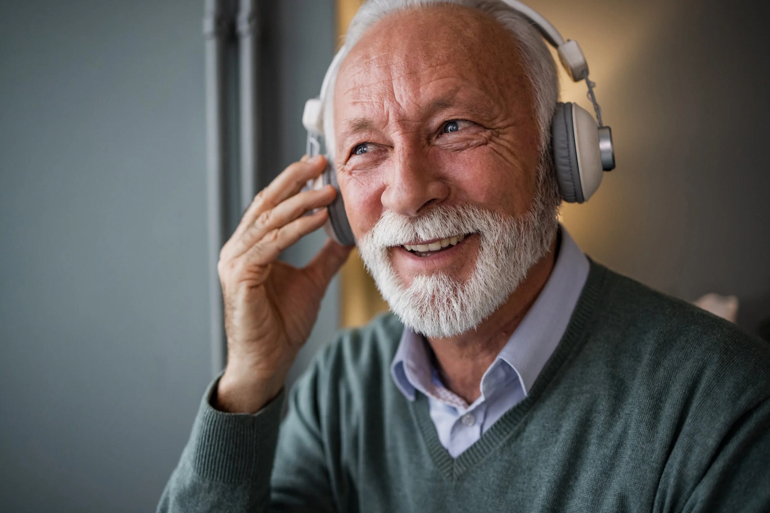 An elderly man with white hair and a beard smiling while wearing large over-ear headphones.