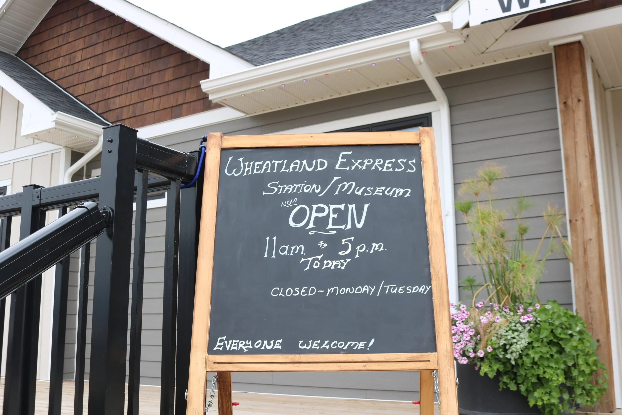 A chalkboard sign outside a building advertising Wheatland Express Station and Museum, open from 11 a.m. to 5 p.m., closed on Monday and Tuesday, with colorful flowers in a pot nearby.