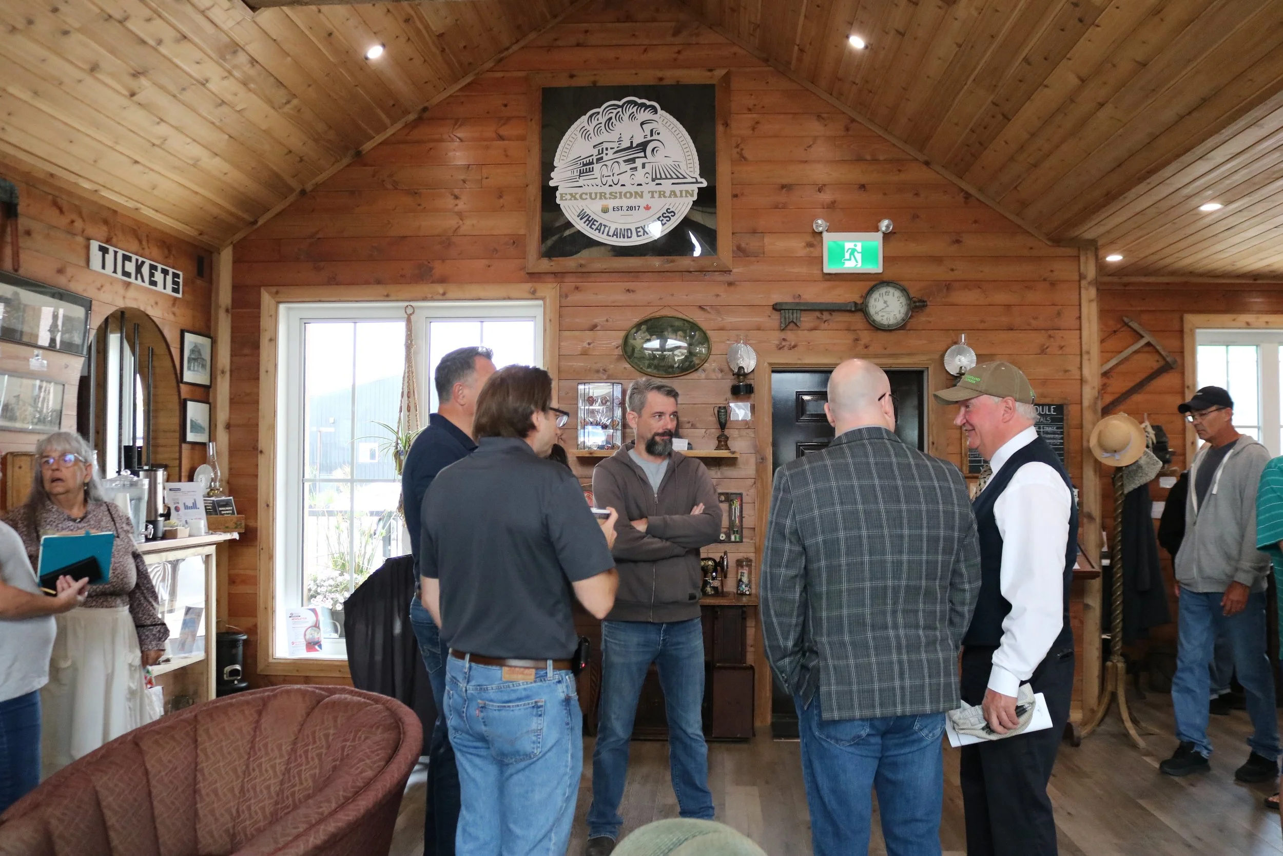 Group of people talking inside a wood-paneled room with a sign that reads 'Tickets' on the wall and a large logo for 'Excursion Train Wheatland Express' above the window.