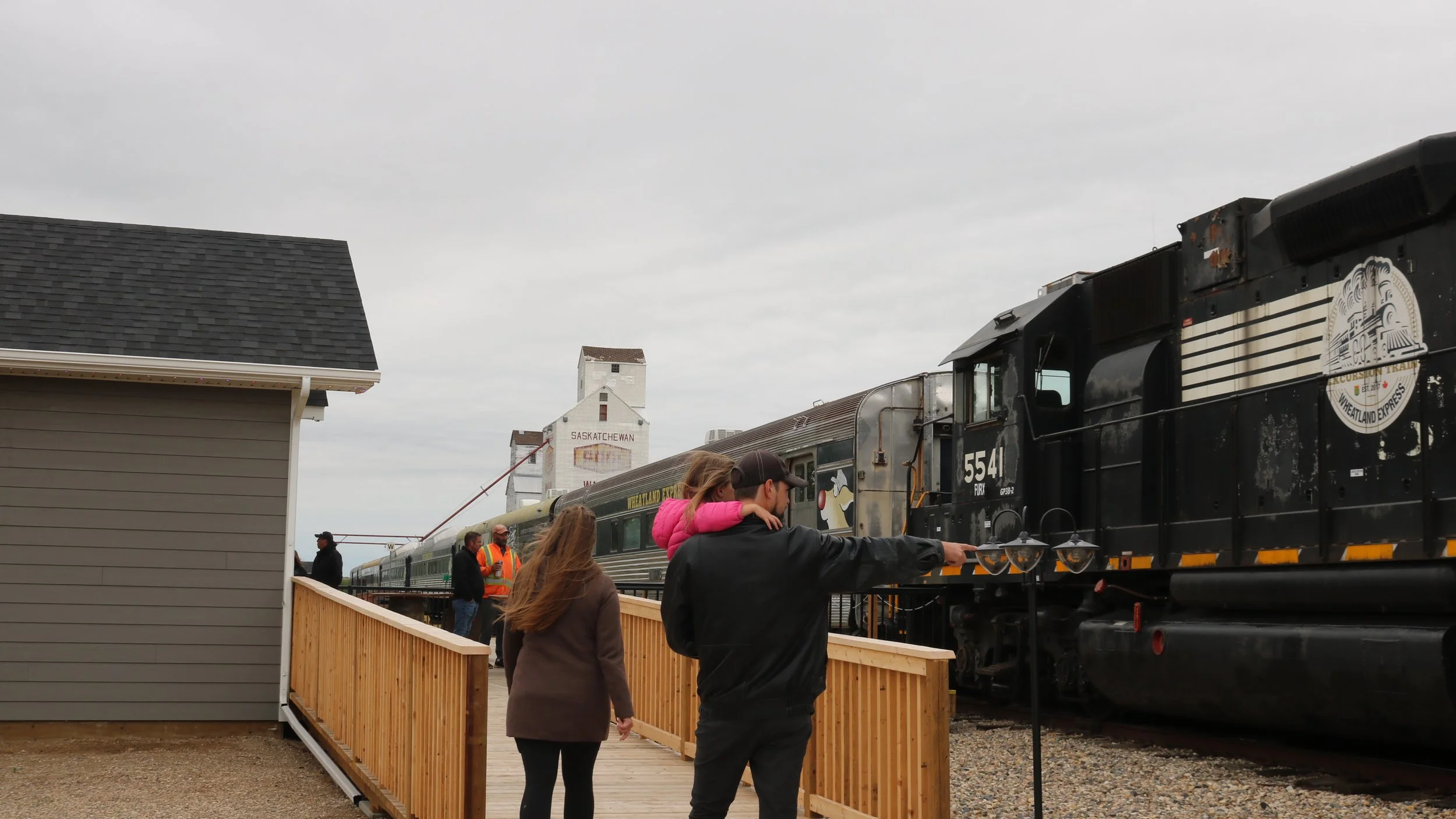 People walking on a wooden platform next to a vintage train with a black locomotive. The scene is outdoors with a cloudy sky and grain elevators in the background.