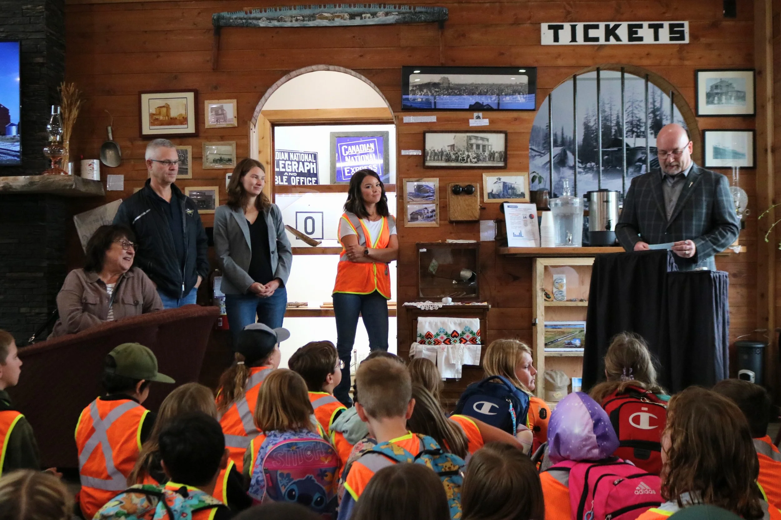 Kids sitting on the floor in orange safety vests inside a wood-paneled room, listening to a man in a suit at a podium and four adults standing behind him near a historical display.