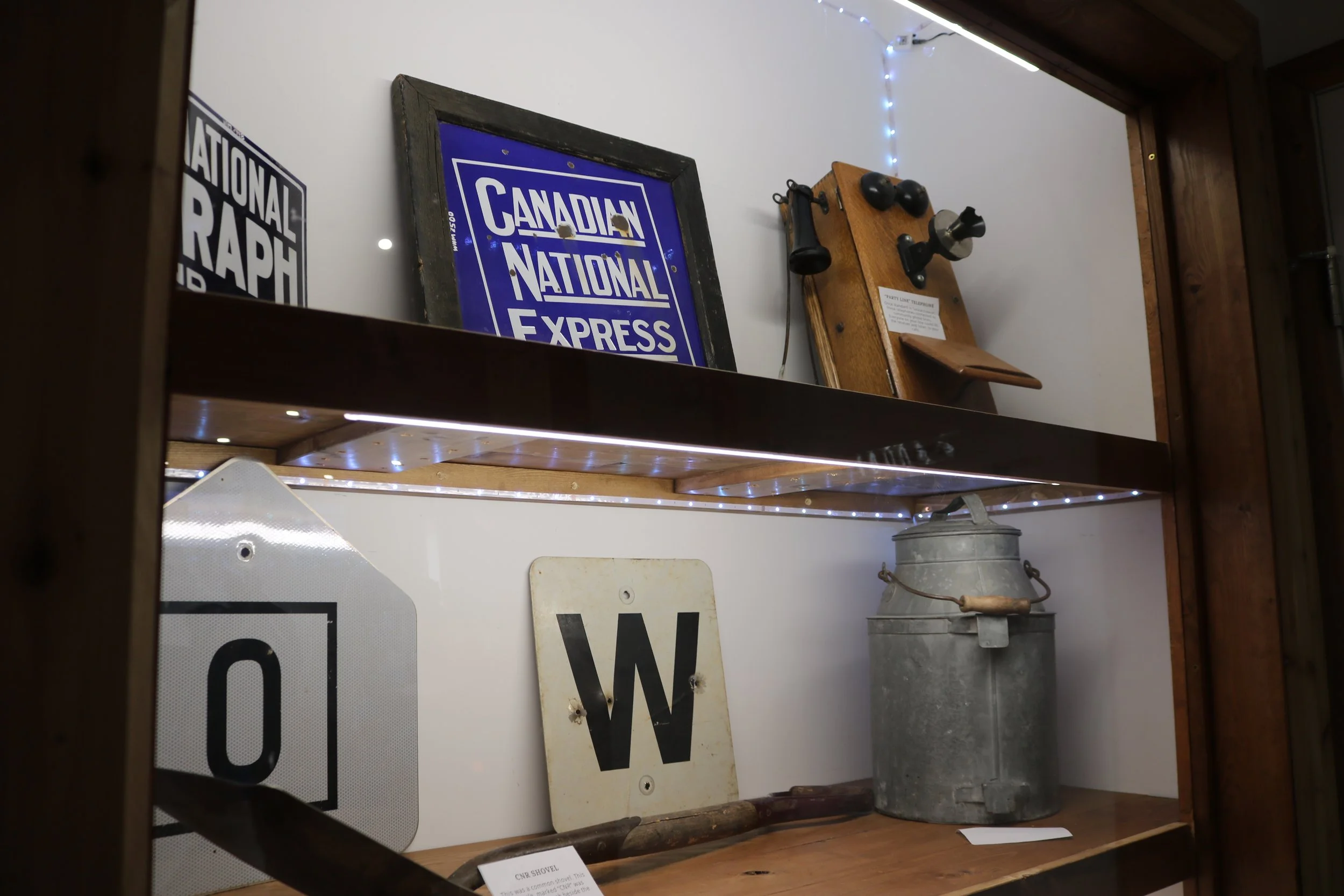 Display case with vintage items including a blue sign reading 'Canadian National Express,' a framed poster, an old wooden wall-mounted telephone, a metal milk can, and railway signs.