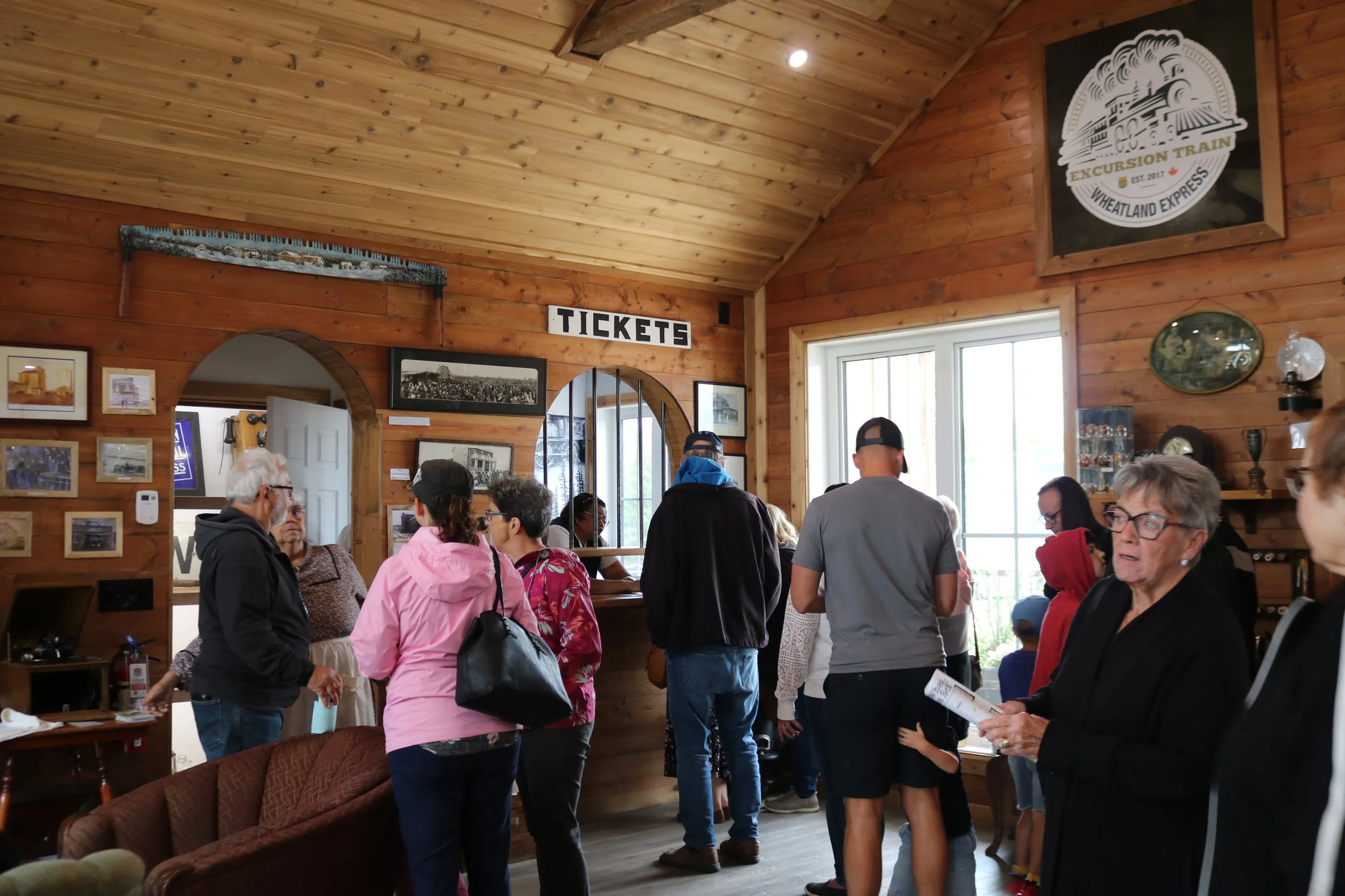 People gathered inside a wooden train station ticket booth, with vintage photos and signs on the wall, and a large window letting in natural light.