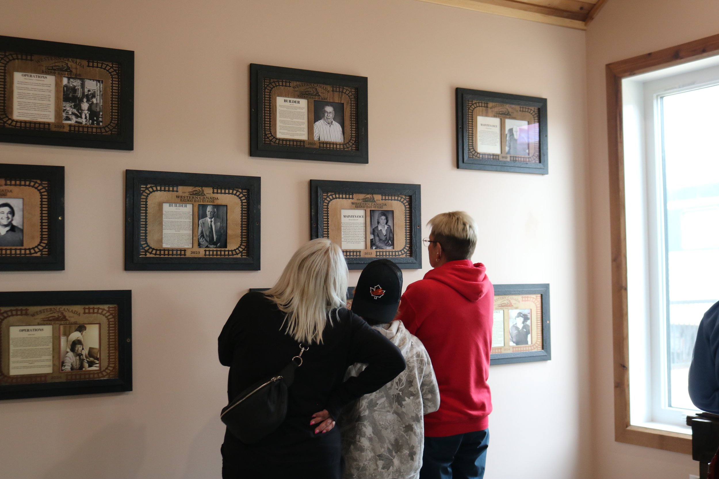 Three people looking at framed photographs and information on a wall in a museum or gallery, near a window.
