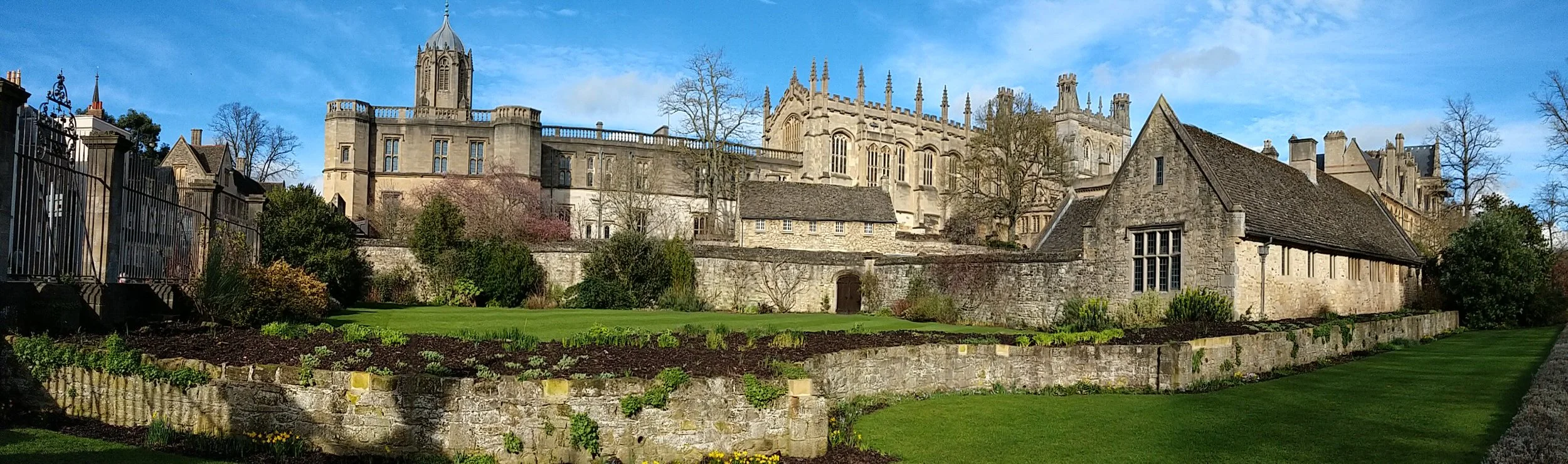 A historic stone castle with tall towers and pointed spires, set on a hill under a partly cloudy blue sky, with a lush green garden in the foreground.