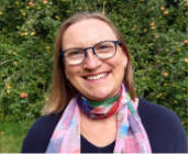 Smiling woman wearing glasses and a colorful scarf outdoors surrounded by green foliage.