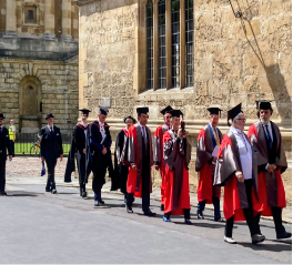 Group of academic graduates in caps and gowns walking outside a historic stone building.