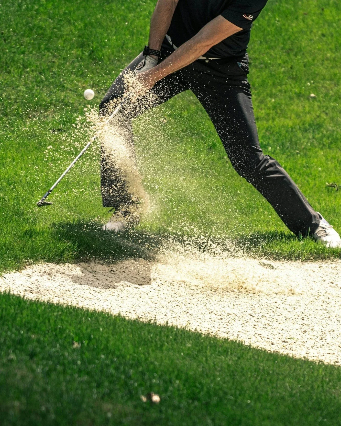 A golfer hitting a golf ball from a sand bunker on a golf course, with sand flying in the air.