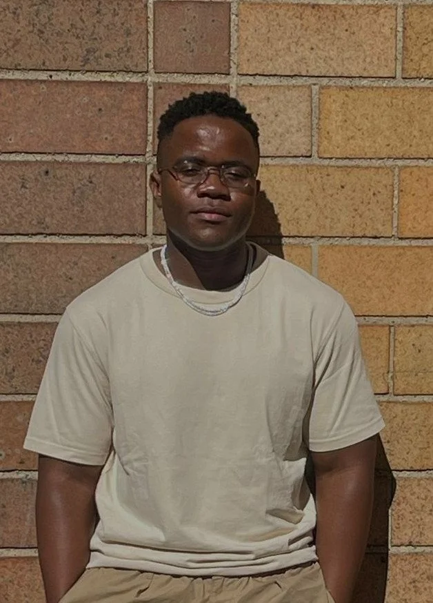 A young man with glasses and short curly hair, wearing a beige T-shirt and necklace, leaning against a brick wall.