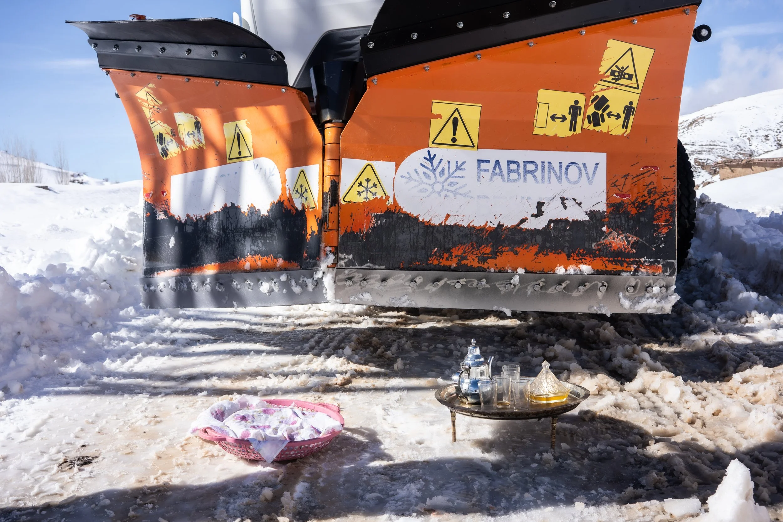 A moment of mountain hospitality set on the snow beneath the snowplow, marking the village’s reconnection to the outer road before the return trip to Imilchil.