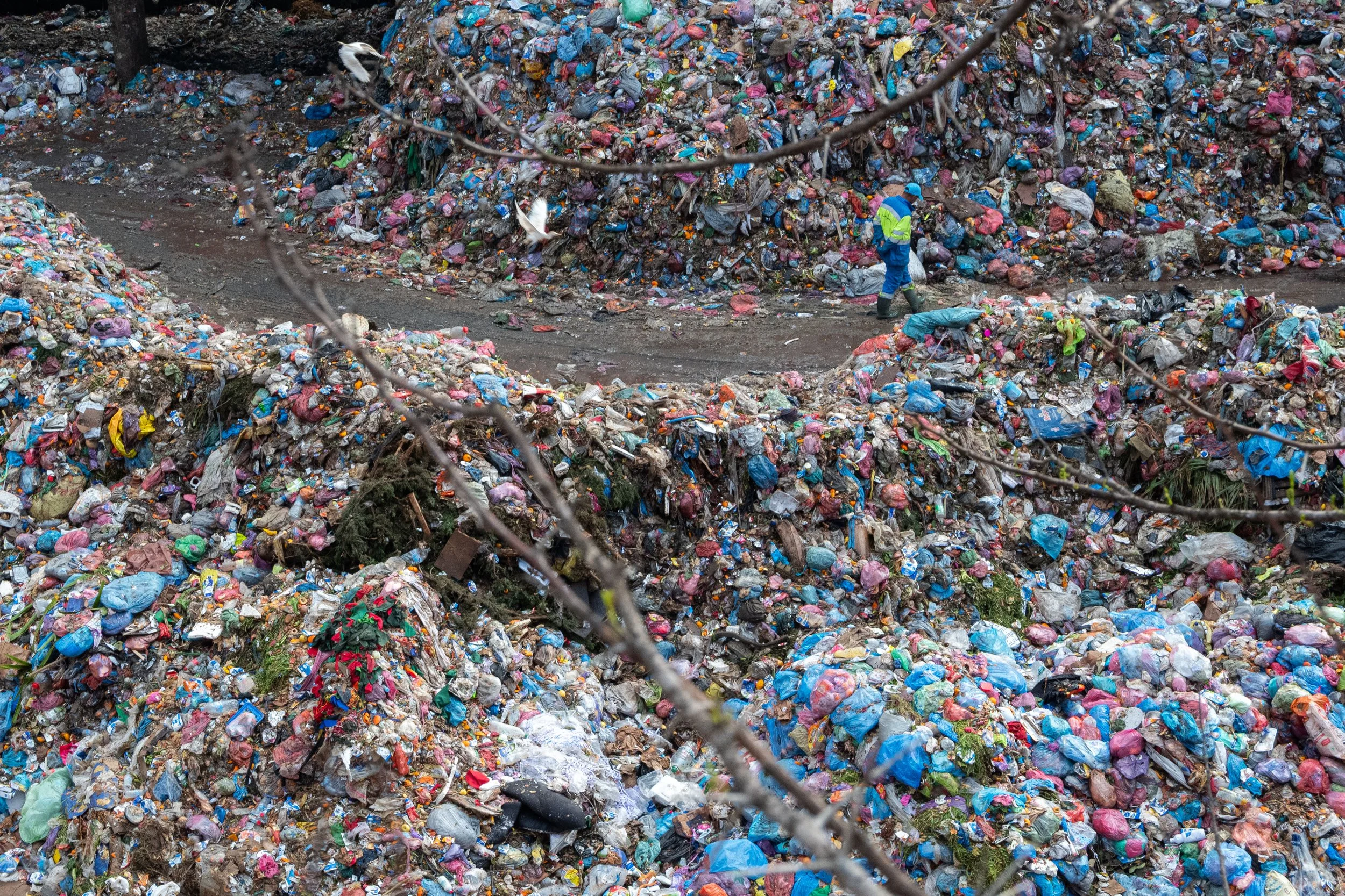 A large landfill filled with assorted trash, including plastic bags and debris, with a worker in a high-visibility jacket and helmet walking along a dirt road through the waste