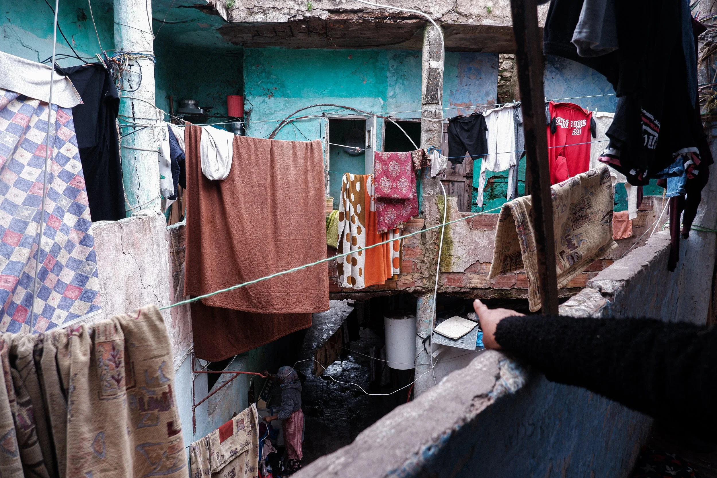 Inside the courtyard of Funduq Hajji, daily life carries on directly alongside the physical collapse of the building. Bright blankets and damp clothes dry on lines tied to crumbling support pillars, even as residents point out sections of the ceiling