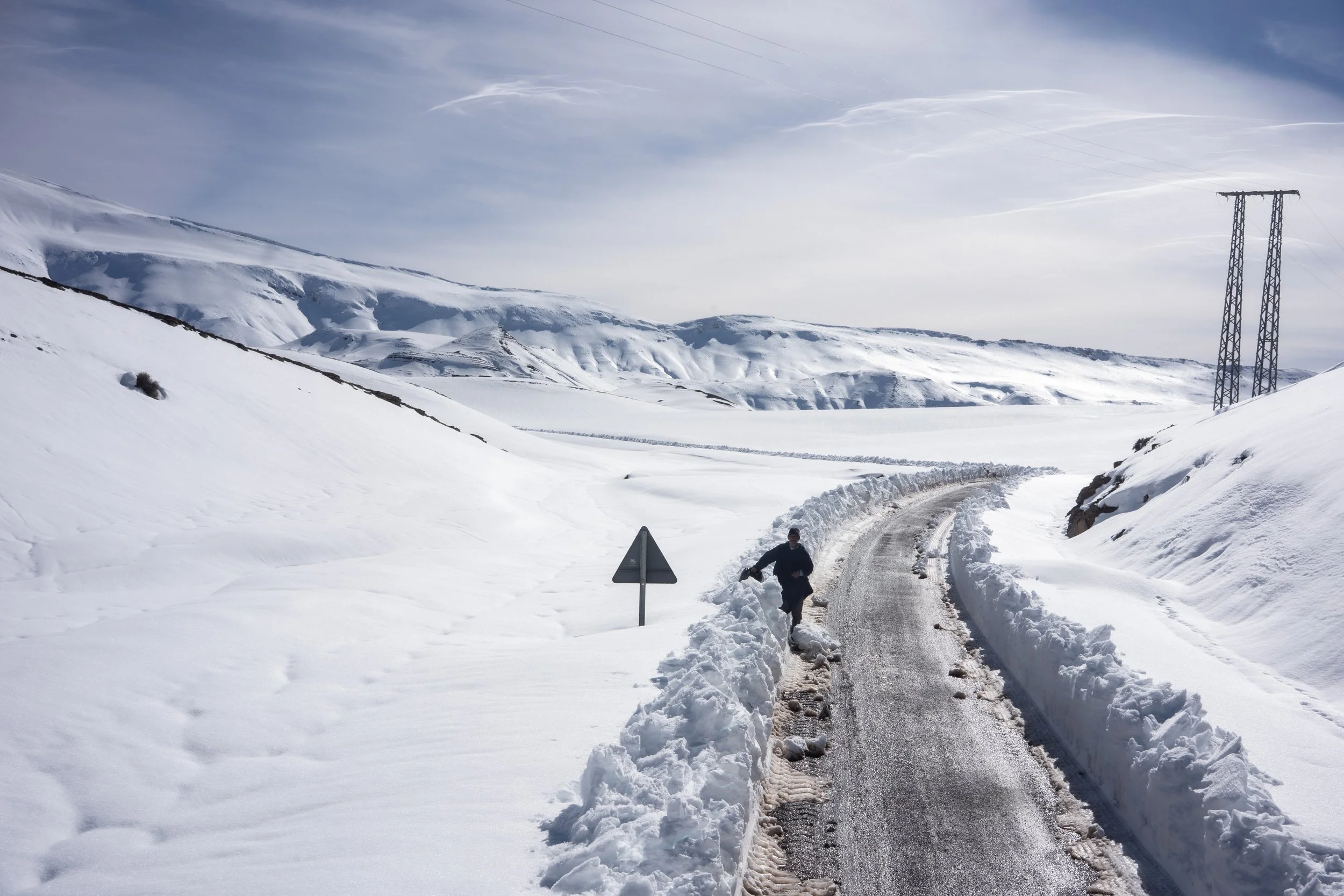 A narrow corridor carved through deep snowbanks reopens travel along the high mountain pass. Walking along the newly plowed ridge, a solitary traveler makes their way along the asphalt path connecting isolated settlements.