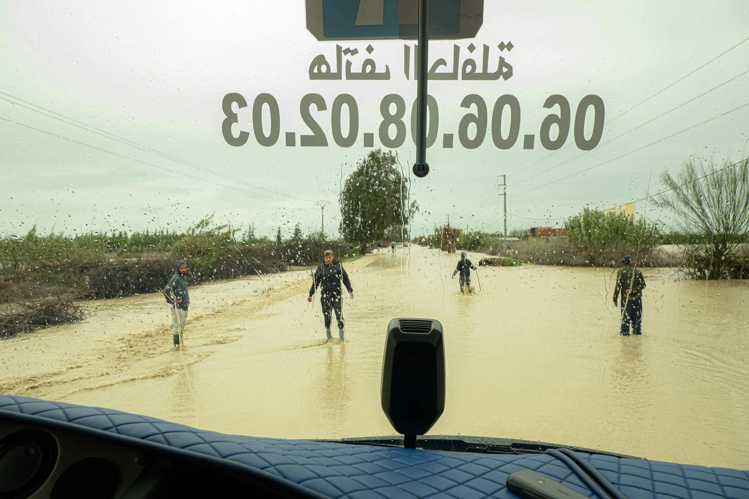 Through the windshield of a stopped vehicle, local spotters test the safety of an inundated transit route. The river overflow has erased visible road boundaries, forcing men to wade ahead on foot to guide traffic through the flooded plain.