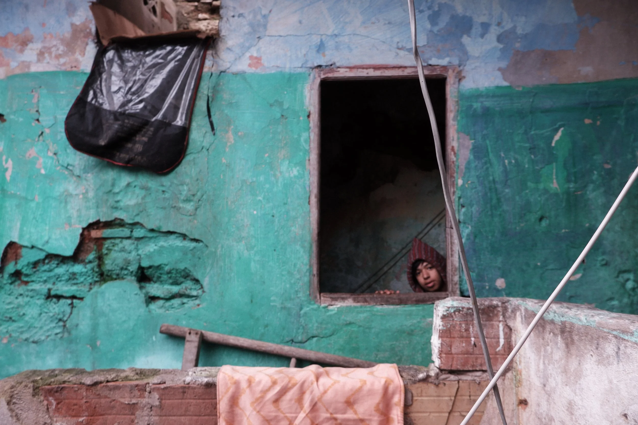 Growing up in a state of suspension. Framed by peeling plaster and cracked masonry, a young face looks out from the shadows of an unstable room, where structural neglect quietly reshapes the boundaries of daily life and home.