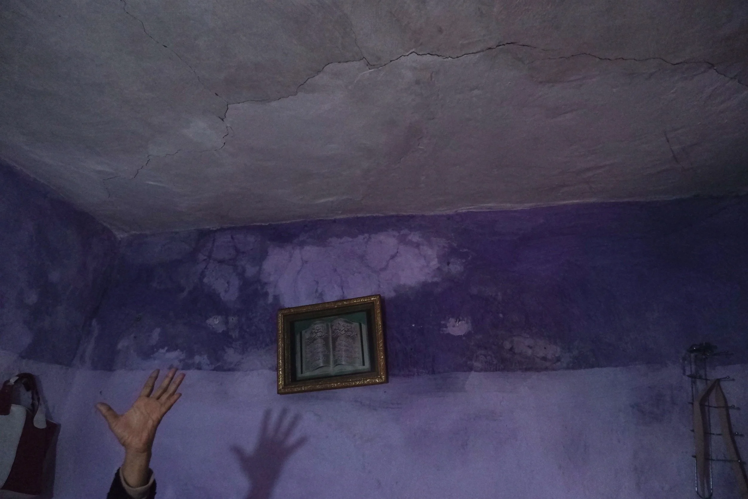 Salé, Morocco. A hand is raised against a damp purple wall, marking the steady progress of a crack splitting the ceiling plaster. In the historic funduqs of Hajji and Korchi, personal fixtures like framed religious art sit alongside severe structural